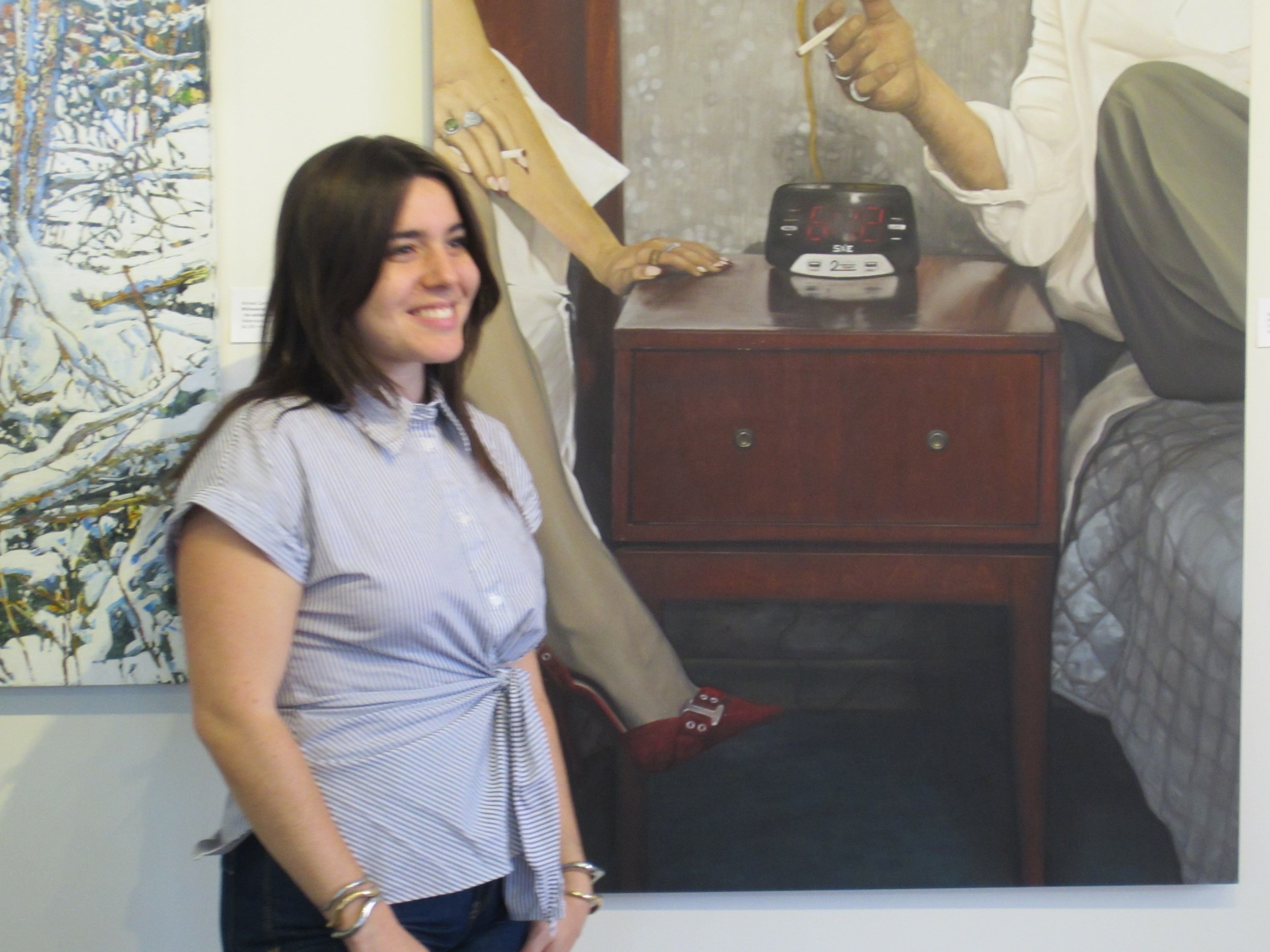 Young woman smiling and standing in front of a painting that depicts a scene with a bed, a woman, a clock on a nightstand, and some artwork on the wall.