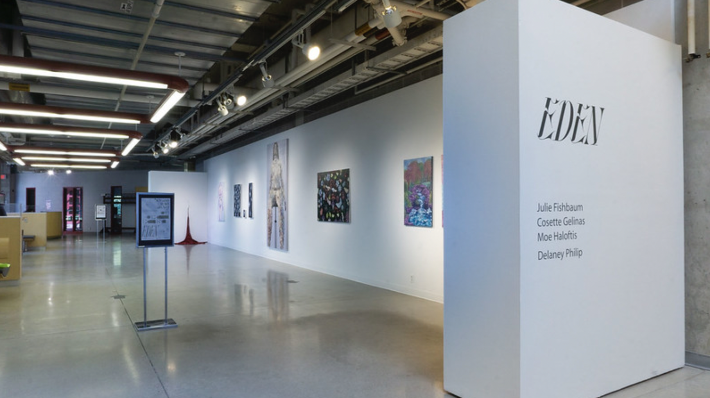 Interior of an art gallery with several paintings displayed on the white walls and informational signs, with a white partition on the right labeled 'EDEN' and a list of artists' names.