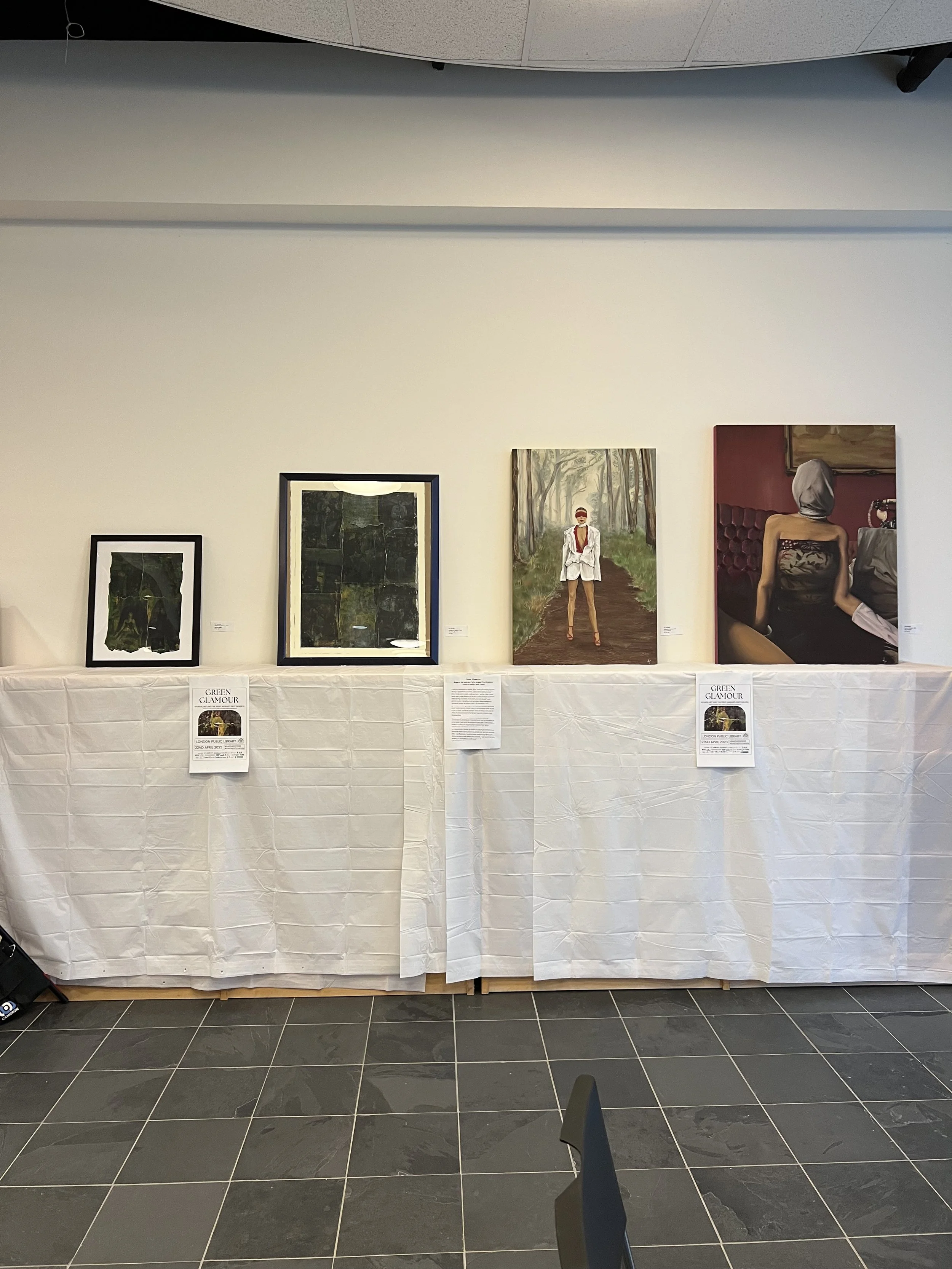 Four paintings displayed on a white tablecloth-covered table at an art exhibition, with two small informational signs underneath.
