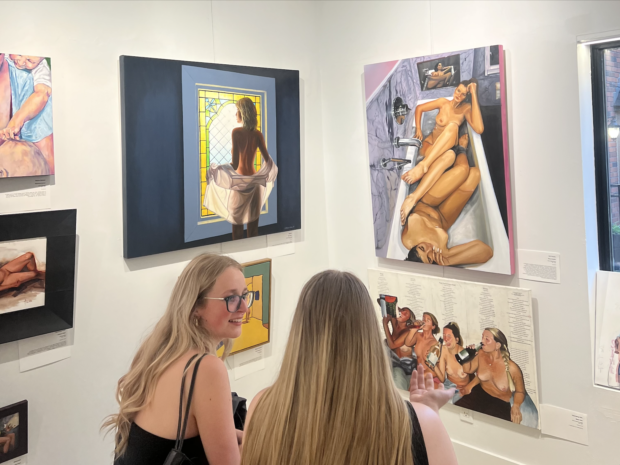 Two women with long blonde hair and glasses viewing paintings of nude women in an art gallery.