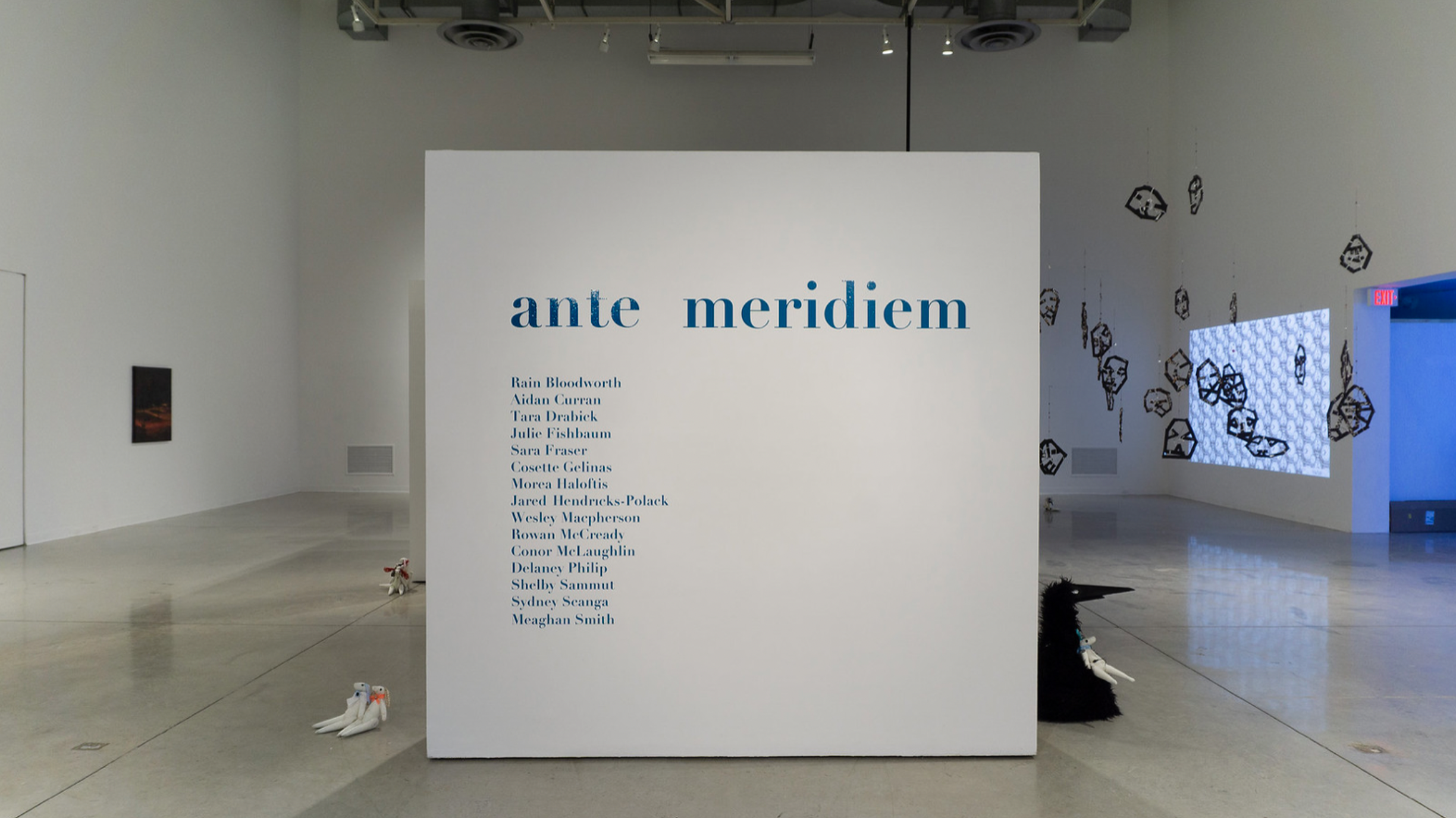 An art exhibit at Ante Meridiem featuring a large white wall with blue text listing artists, in a spacious gallery with artworks on the walls and plush toys on the floor.
