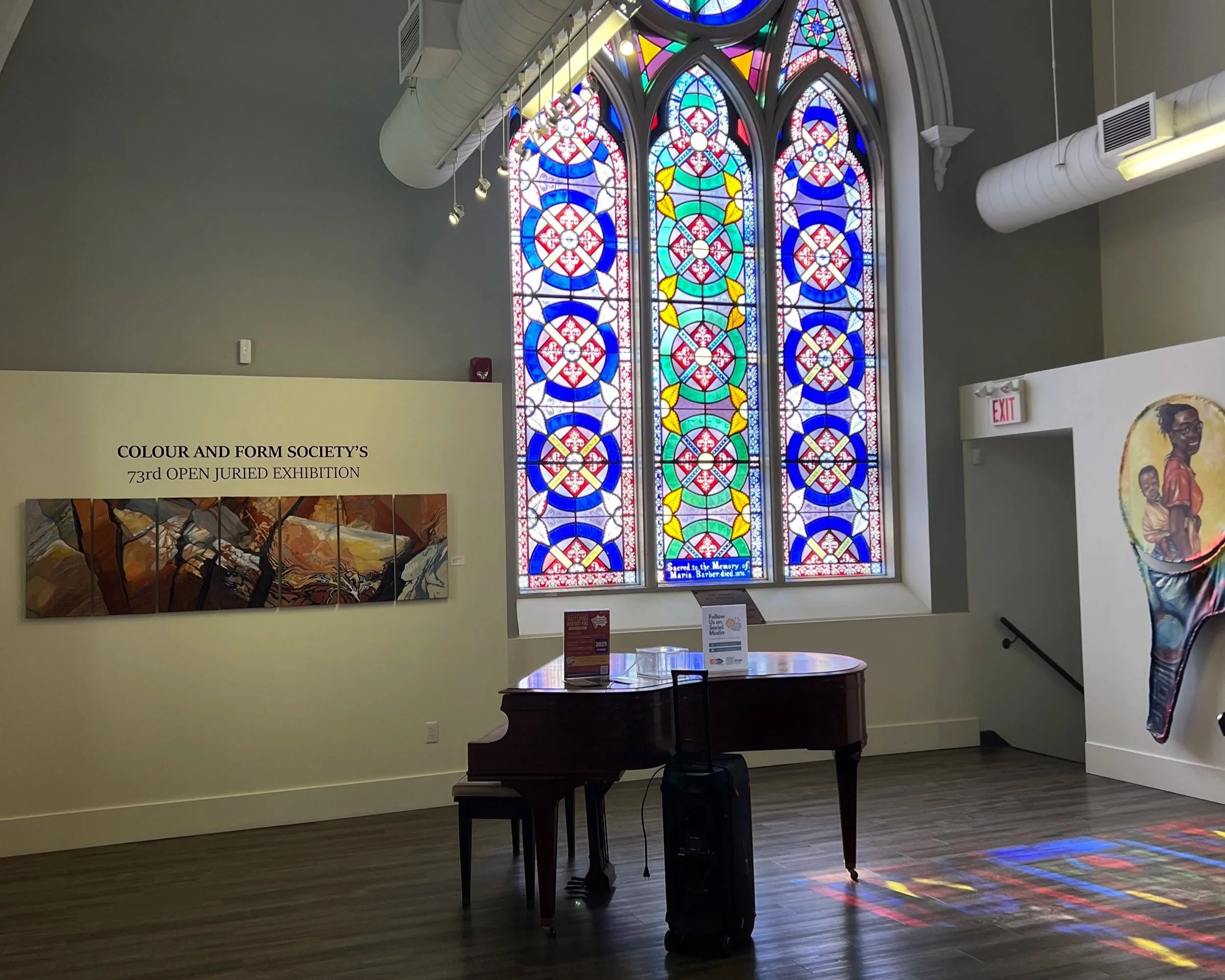 Interior of an art gallery with a large stained glass window, a grand piano, and artworks on the walls. The stained glass includes intricate colorful patterns. A sign announces the 'Color and Form Society's 73rd Open Juried Exhibition'.