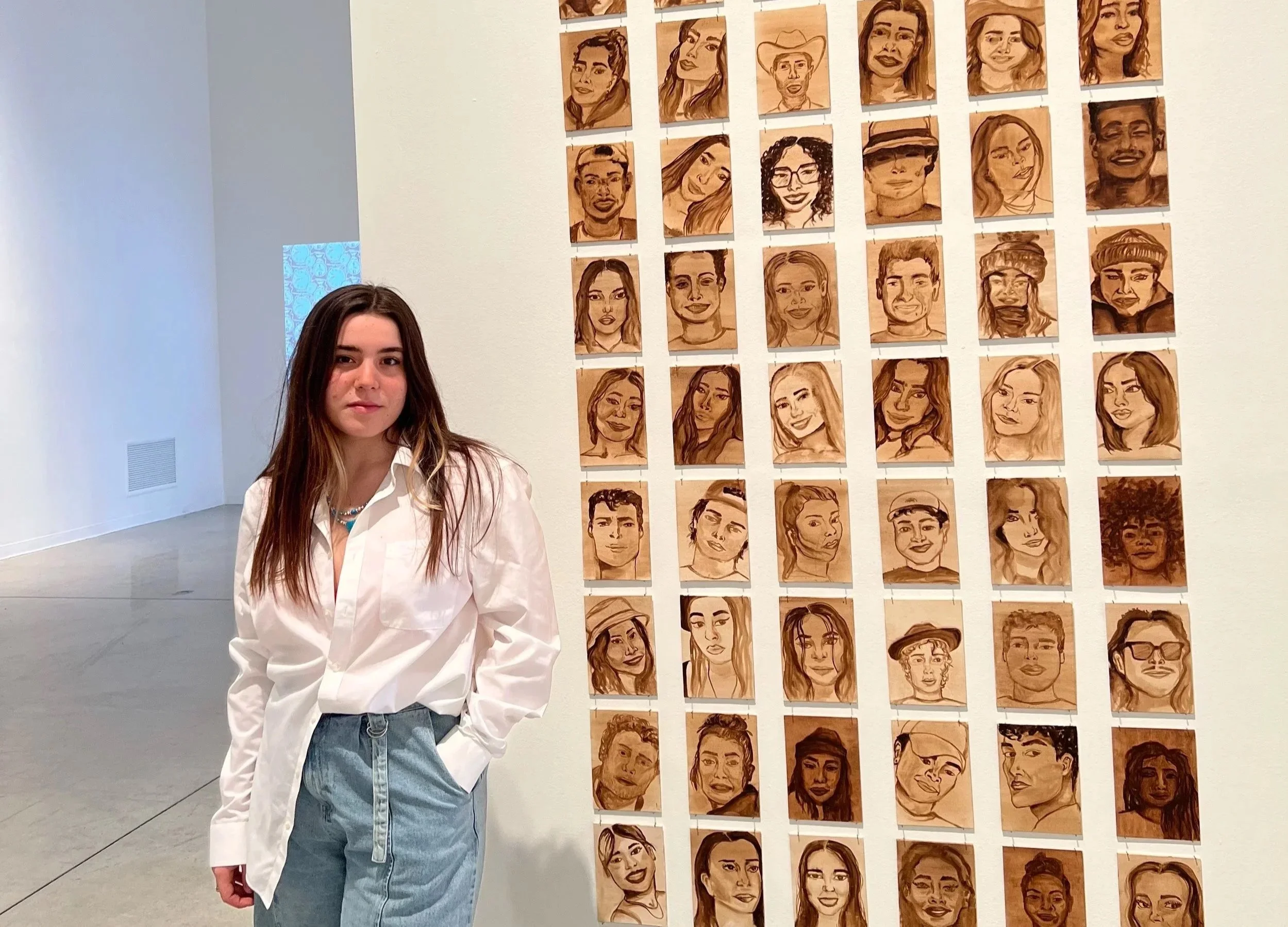 A young woman with long brown hair, wearing a white shirt, light blue jeans, and a pearl necklace, standing with hands in pockets next to a wall display of many portrait sketches on wood panels in an art gallery.