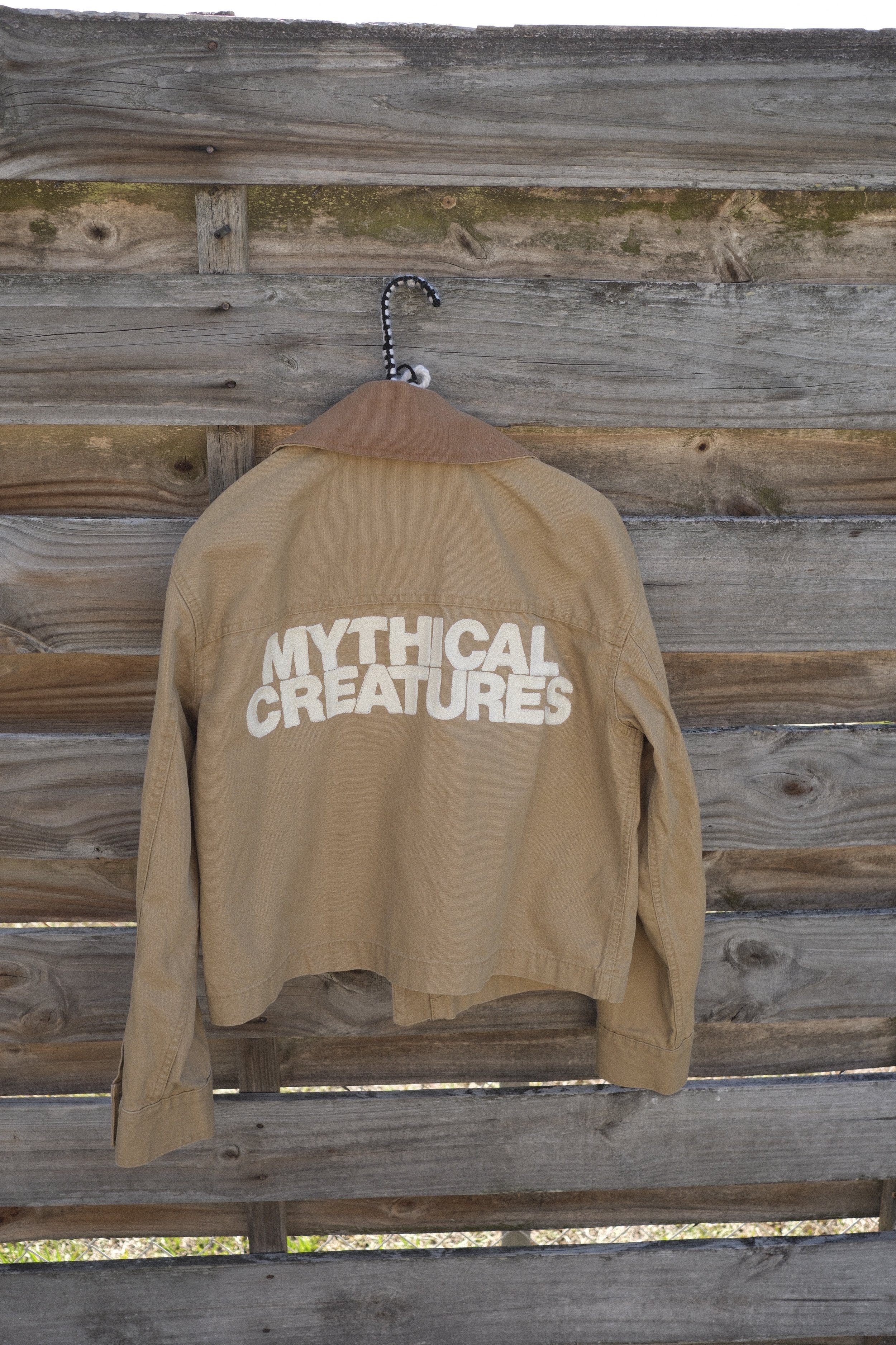 A tan canvas jacket with cream white chainstitch embroidery on the back reading "Mythical Creatures"