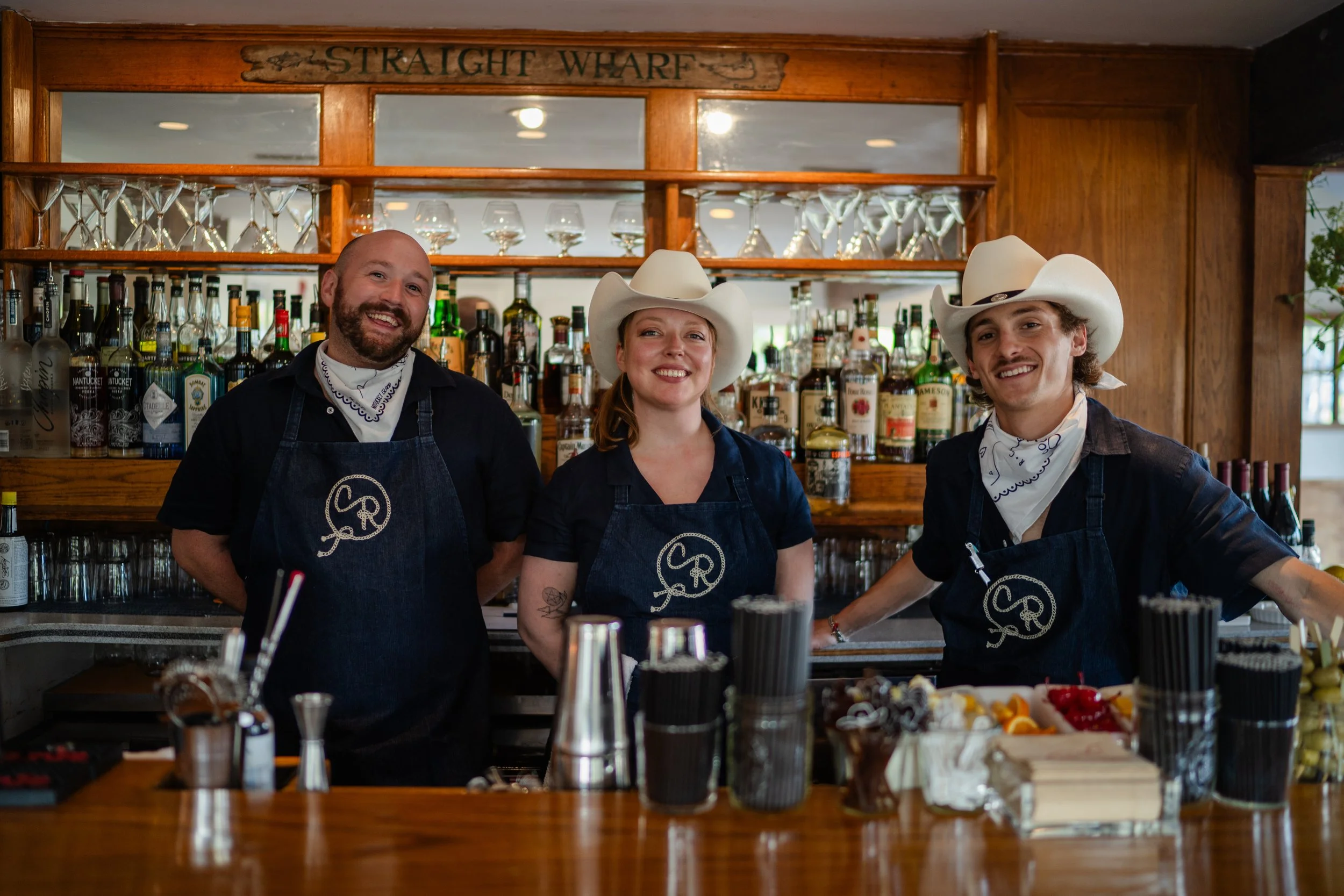 Three people wearing western wear and custom chainstitched aprons with CR stitched in a lasso style, standing behind a bar.
