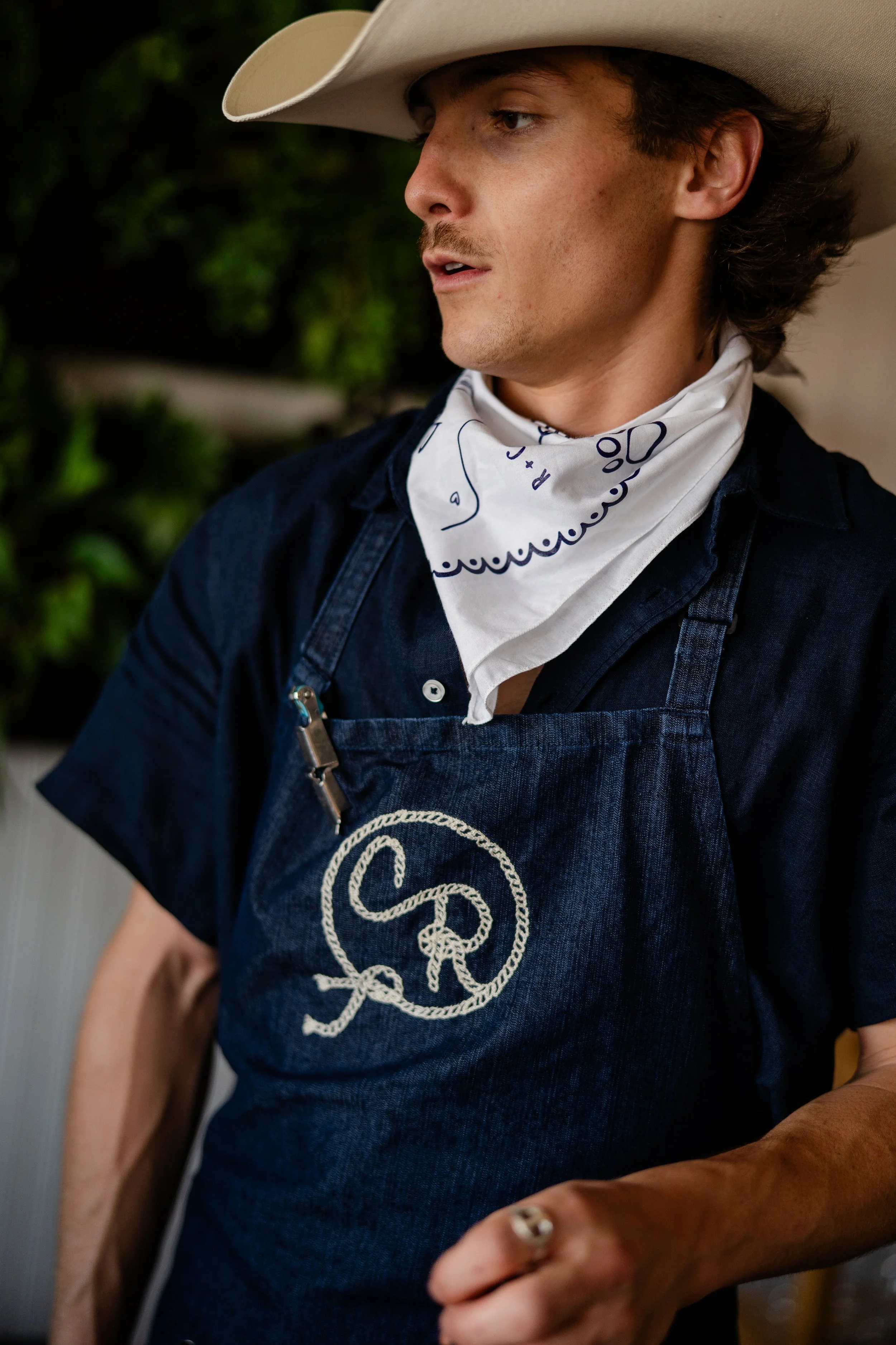 A man in a cowboy hat wearing a custom bandana and chainstitch embroidered denim apron. The logo on the apron is a C and R looped together to look like a lasso.