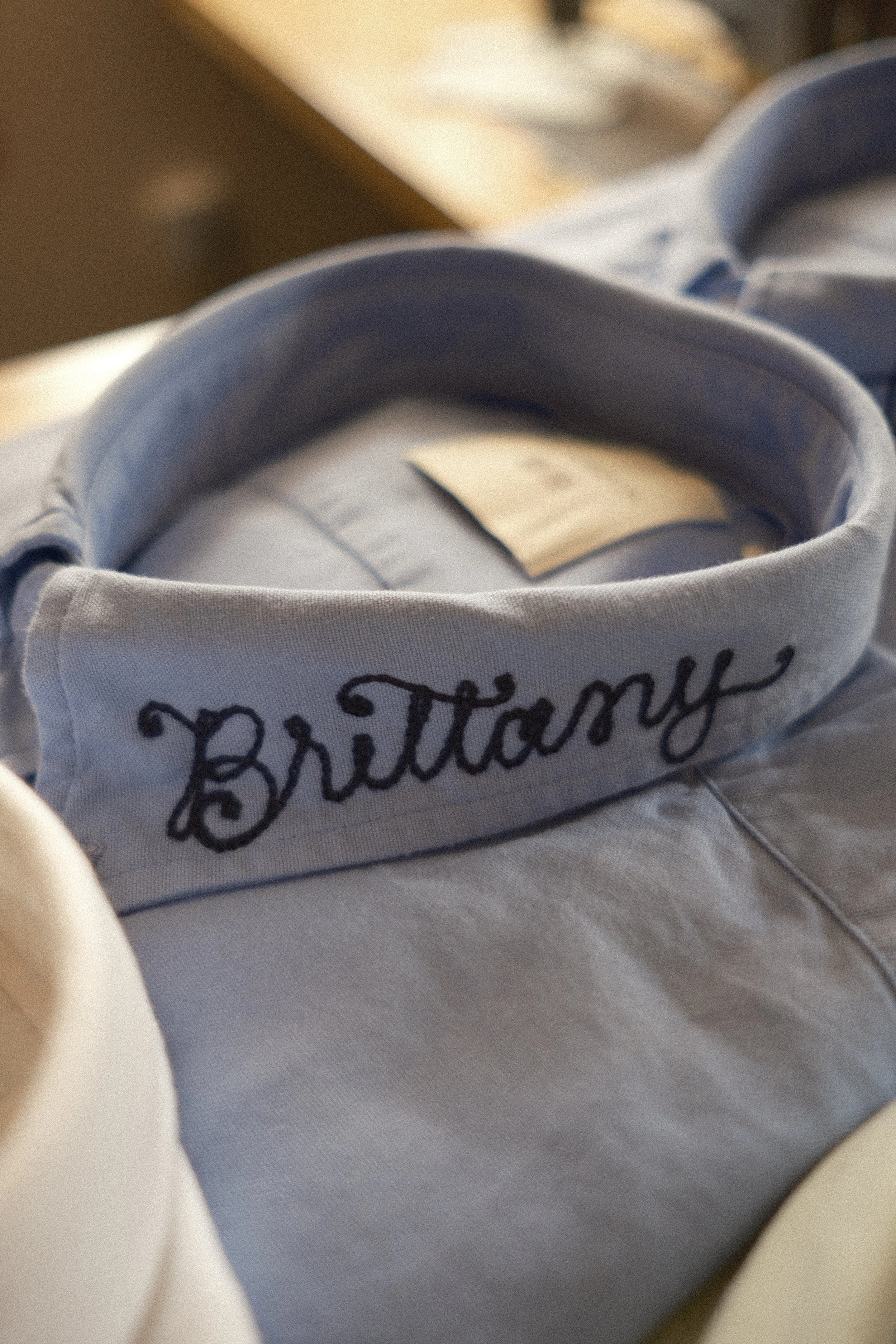 A light blue men's collared dress shirt with "Brittany" in navy blue chainstitch embroidery