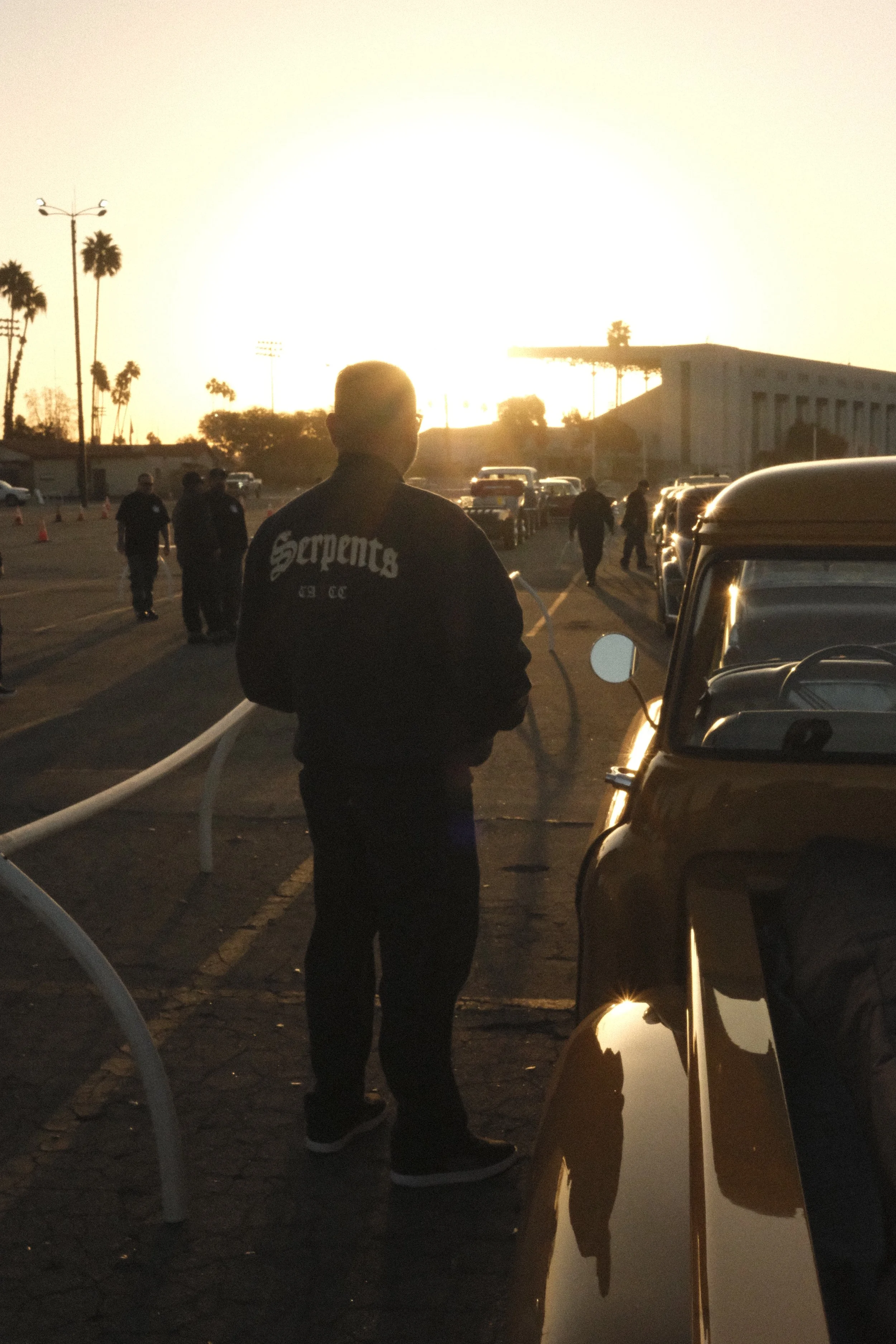 A man standing in a parking lot next to a classic pickup truck, facing the sun wearing a chainstitch jacket with the lettering "Serpents" car club on the back