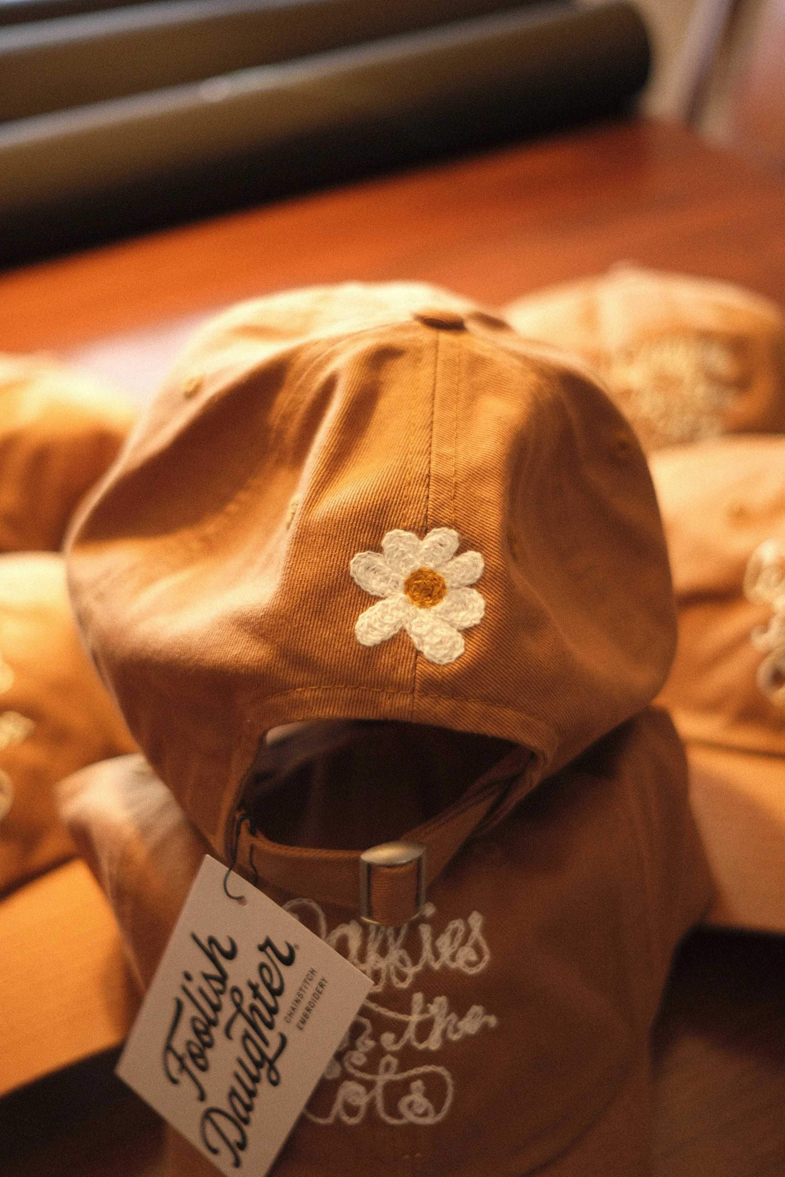 A stack of golden yellow hats, the bottom reading "Daffies in the Cots" in white chainstitch script, and the top hat showing a chainstitch white daisy with yellow center on the back