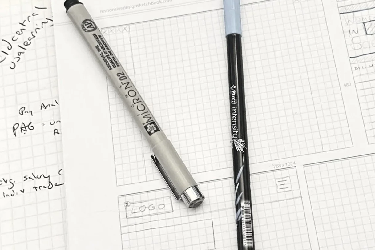 A white sheet of graph paper with the word 'LOGO' handwritten on it, a silver Sharpie marker, and a black ink pen, all placed on a larger sheet of handwritten notes and sketches.