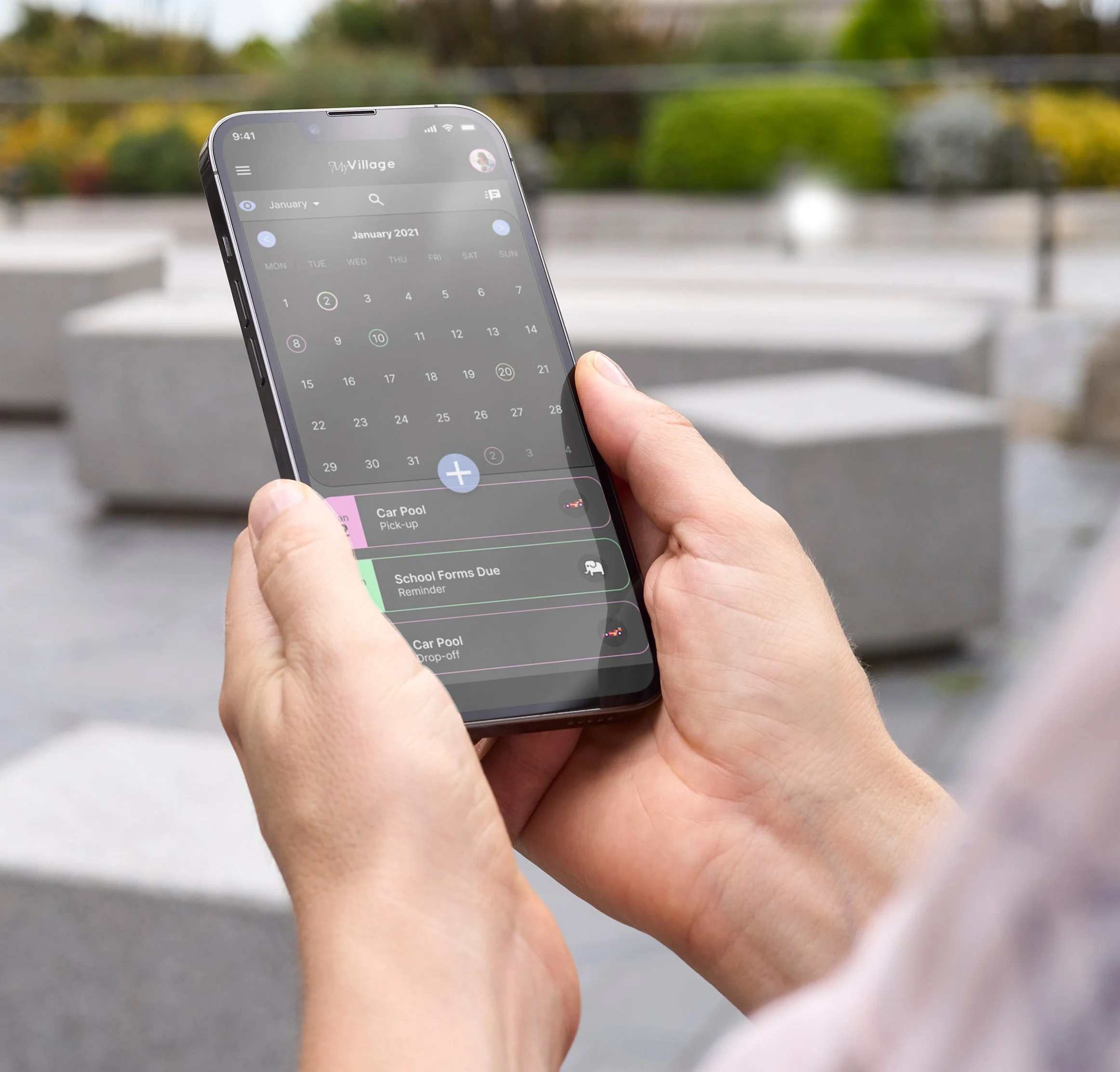 Person holding a smartphone displaying a calendar app with scheduled events, outdoors in a public space with large concrete planters and greenery in the background.