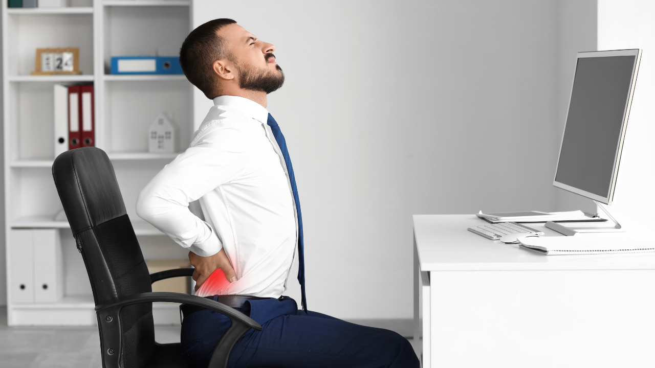 A professional in his mid-40s sitting at a desk in a San Francisco office with visible tension in his hips and lower back, representing the physical cost of prolonged sitting.