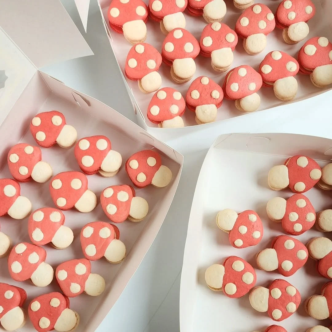 Tray of mushroom-shaped French macarons with red caps and white polka dots, and white stems, arranged in three white boxes.