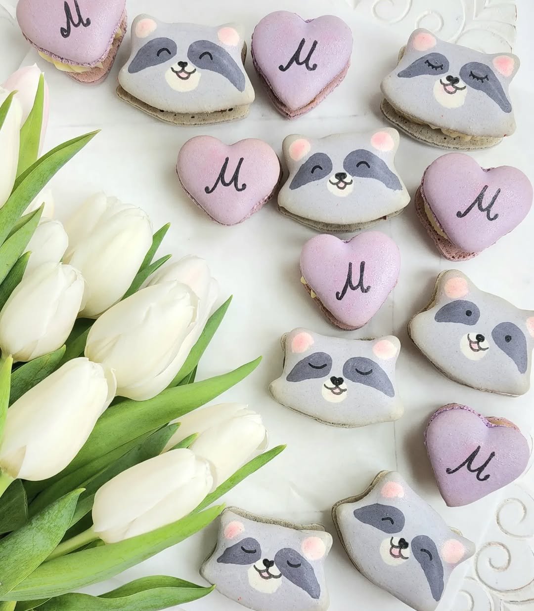 Decorative specialty macarons with raccoon and heart shapes, featuring pastel colors and the letter M, surrounded by white tulips on a white surface.