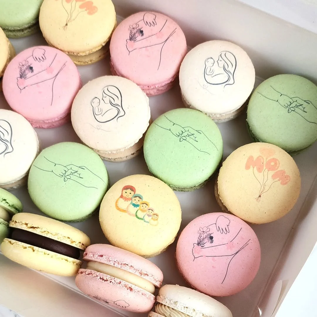Colorful macarons with custom printed illustrations for a baby shower. Graphics include a mother holding a flower, a mother with a baby, a family with children, and hand-holding symbols.