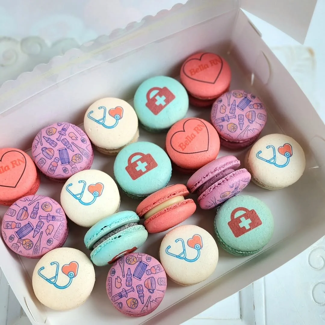 Colorful specialty macarons decorated with medical-themed symbols like stethoscopes, syringes, and first aid kits in a white box.