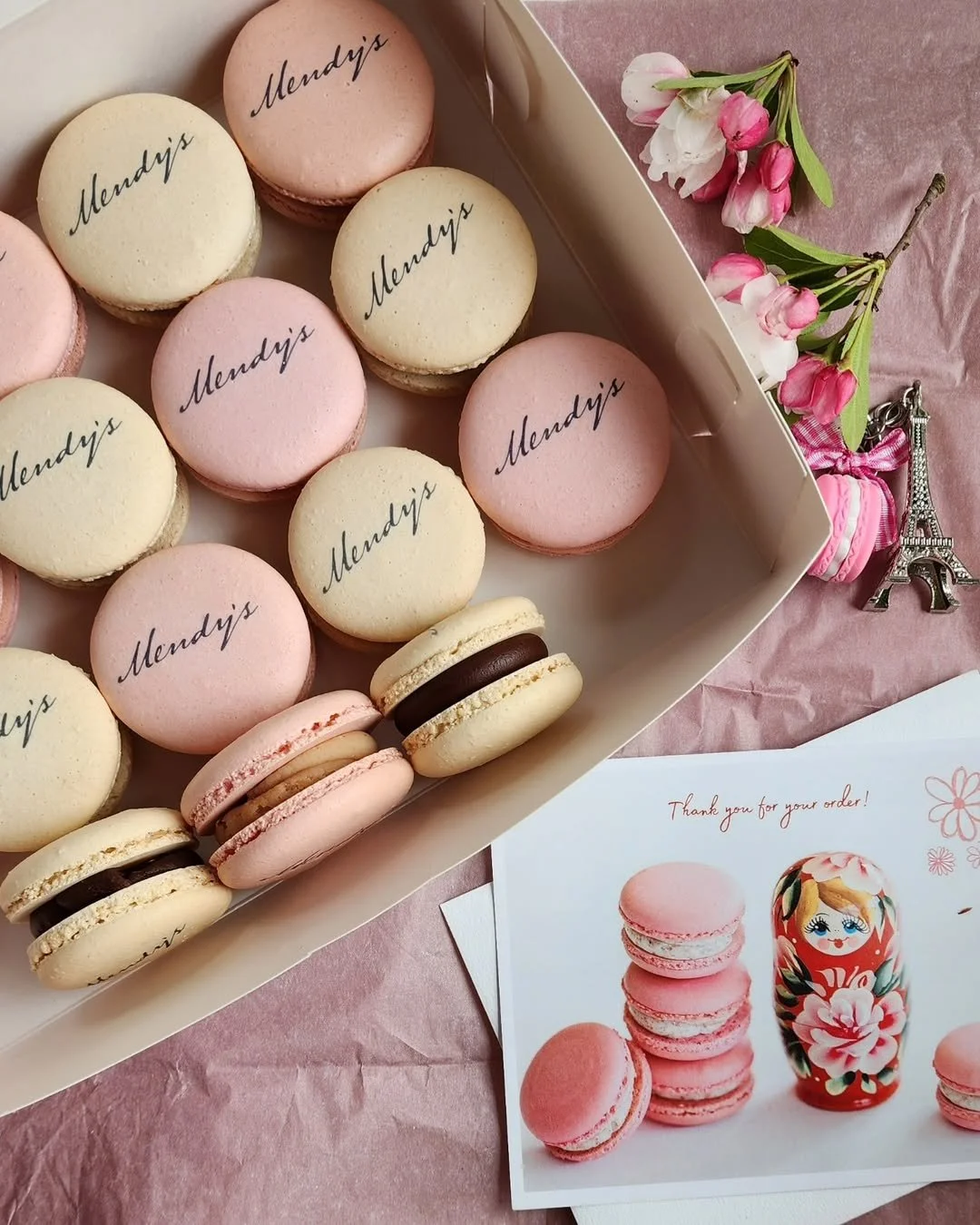 A box of pink and cream macarons with the word 'Mendy's' written on top, placed on pink tissue paper. Next to the box, there are pink and white flowers, a small Eiffel Tower figurine, and a card with a stack of macarons and a Russian nesting doll ill