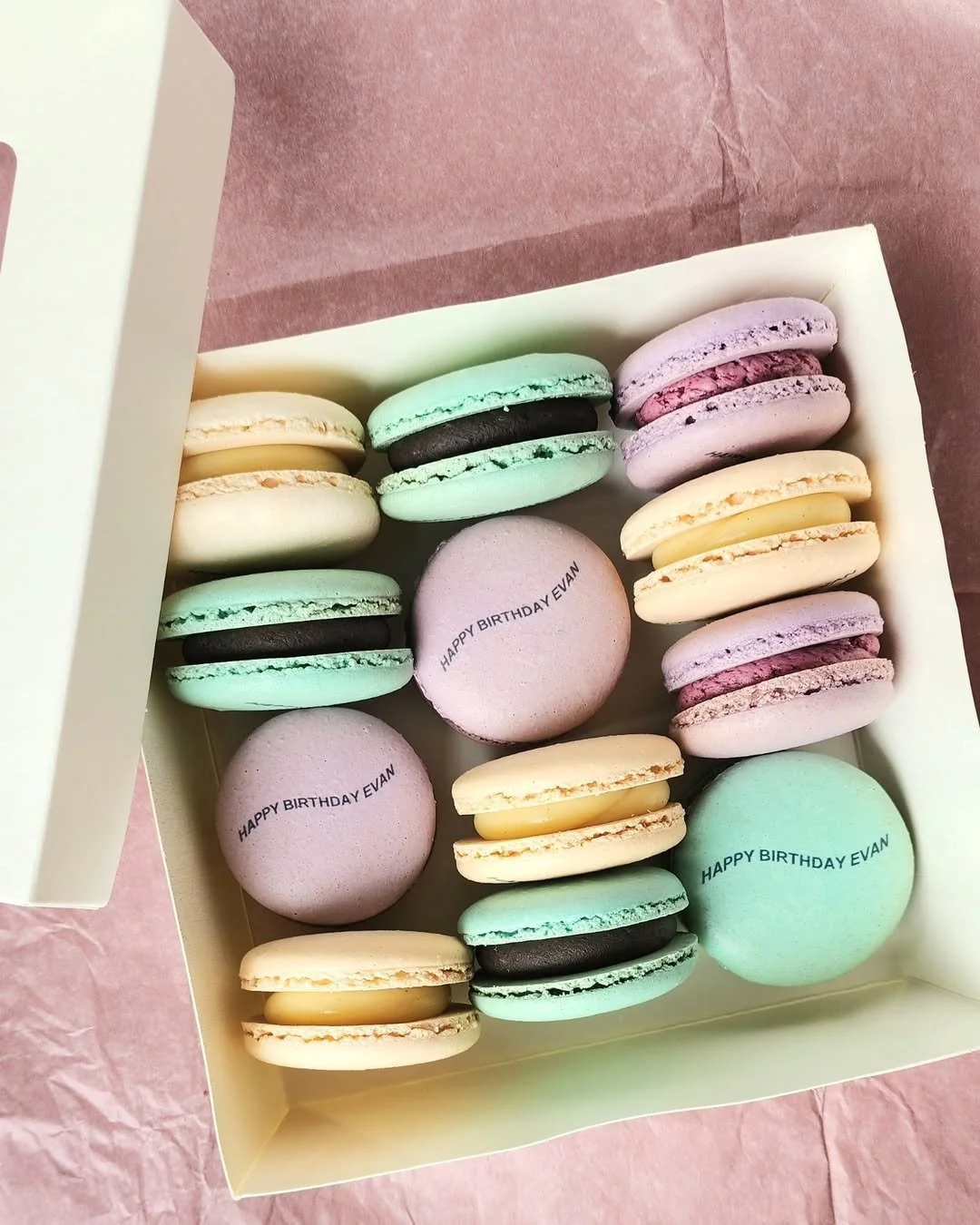 Colorful specialty macarons in a box, each with the message "Happy Birthday Evan" printed on them, in pastel shades of pink, purple, yellow, green, and black, on pink tissue paper.