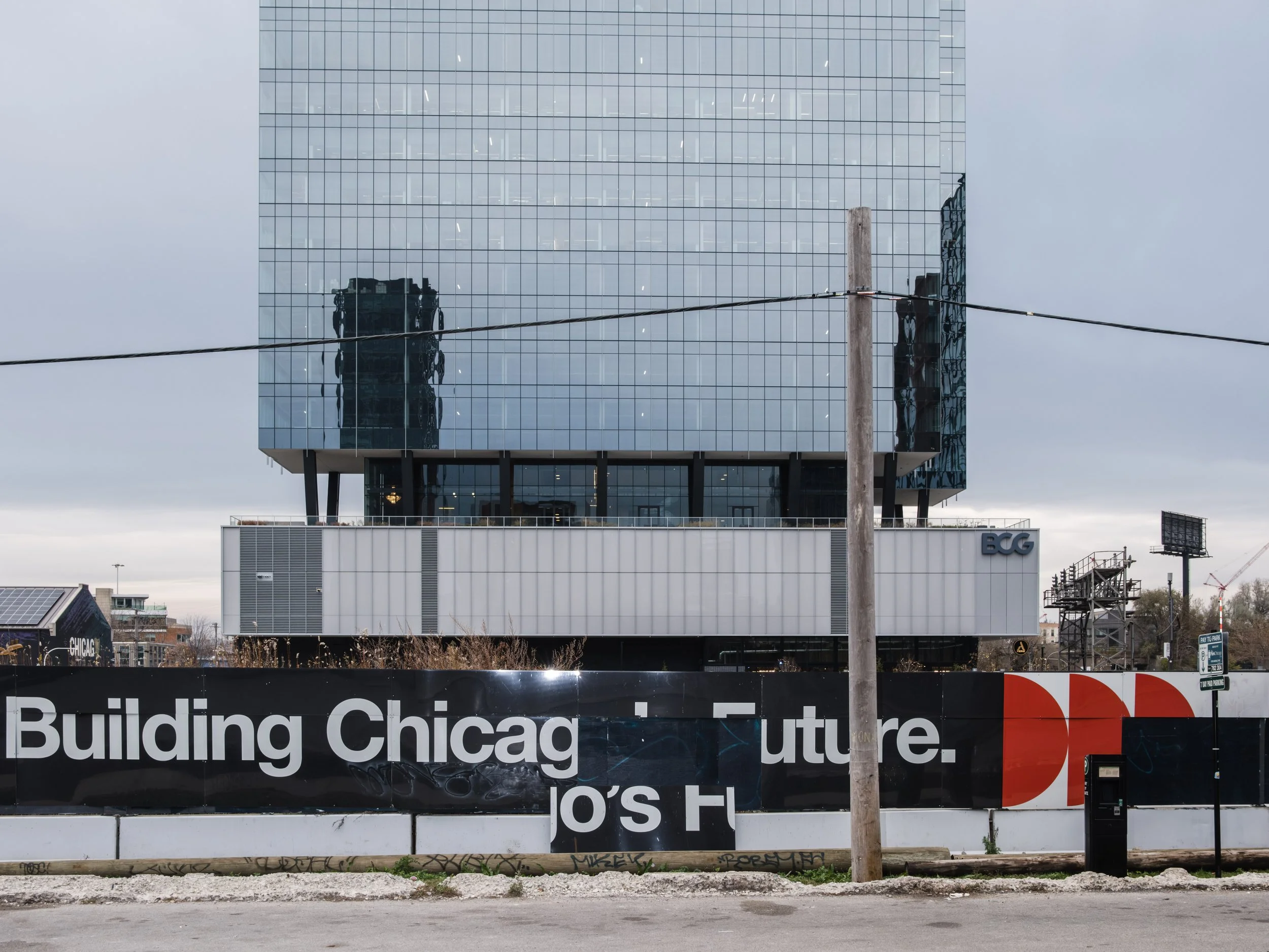 Construction site featuring a modern glass building with a billboard that reads "Building Chicago Future" in front, and a wooden utility pole and a parking meter in the foreground.