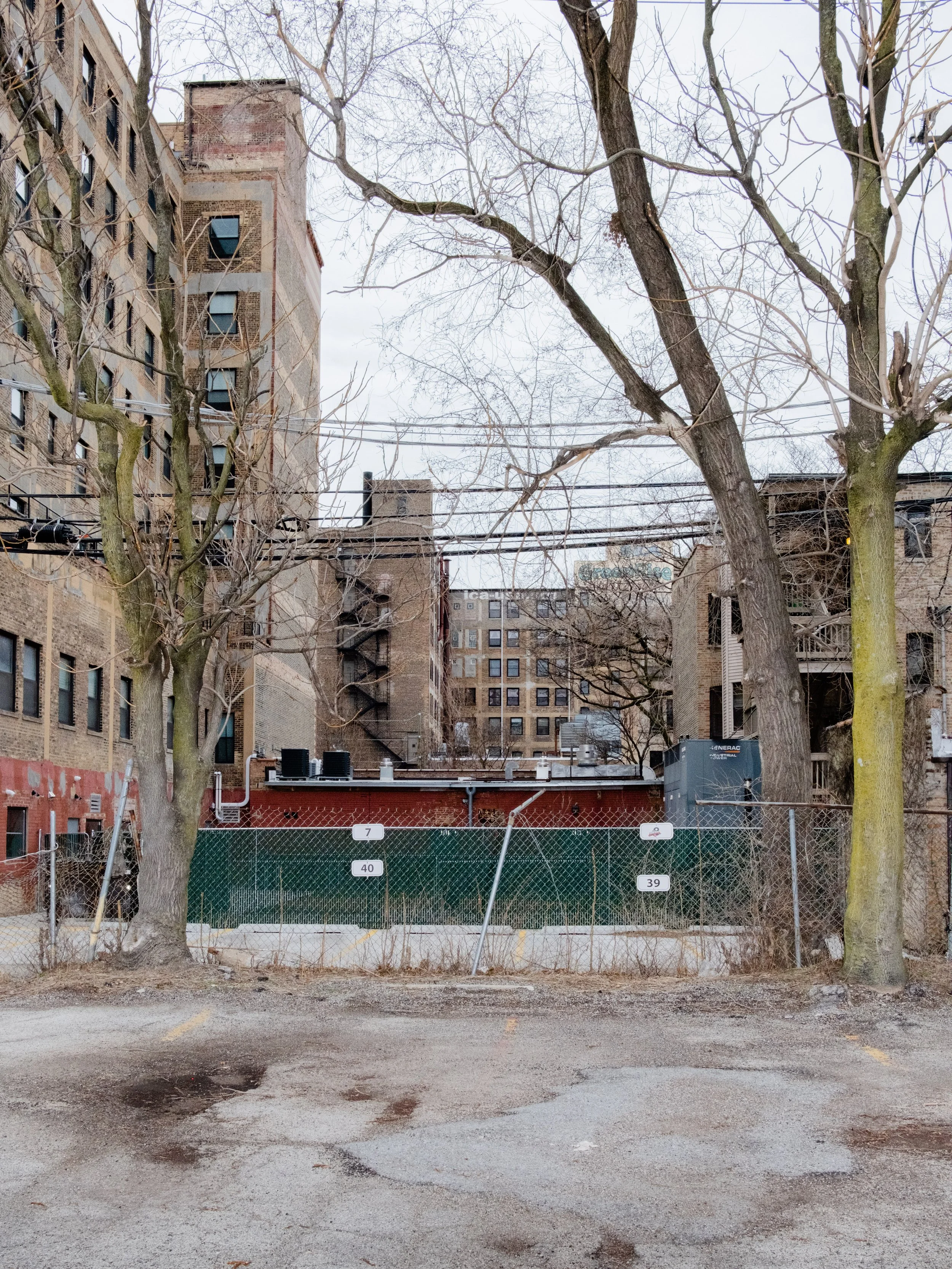 Urban scene showing leafless trees, a parking lot with damaged asphalt, a chain-link fence, and multiple multi-story brick buildings in the background.