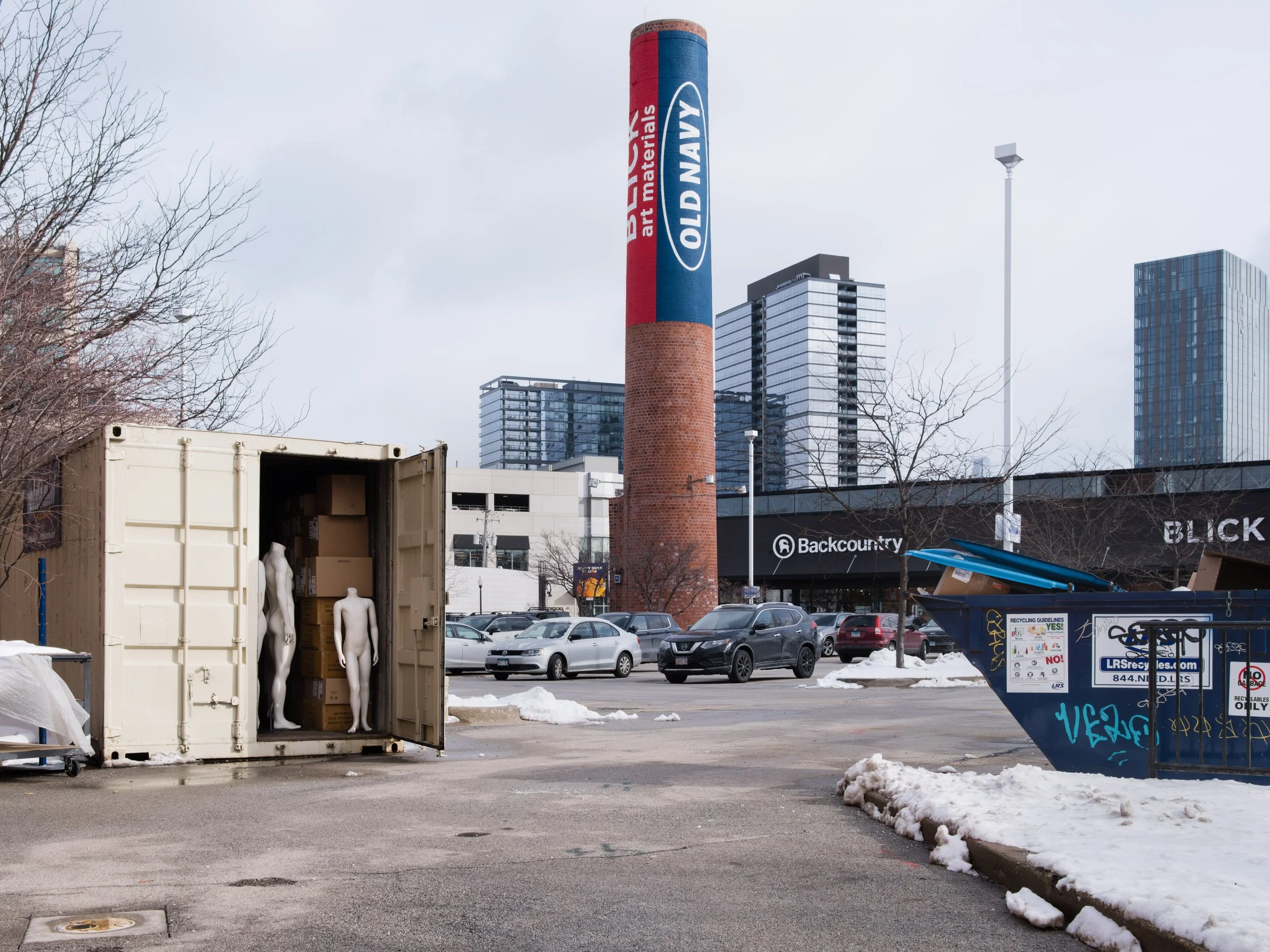 An outdoor parking lot with a shipping container containing two mannequins and boxes, a blue dumpster, surrounding cars, snow patches, and modern high-rise buildings in the background.