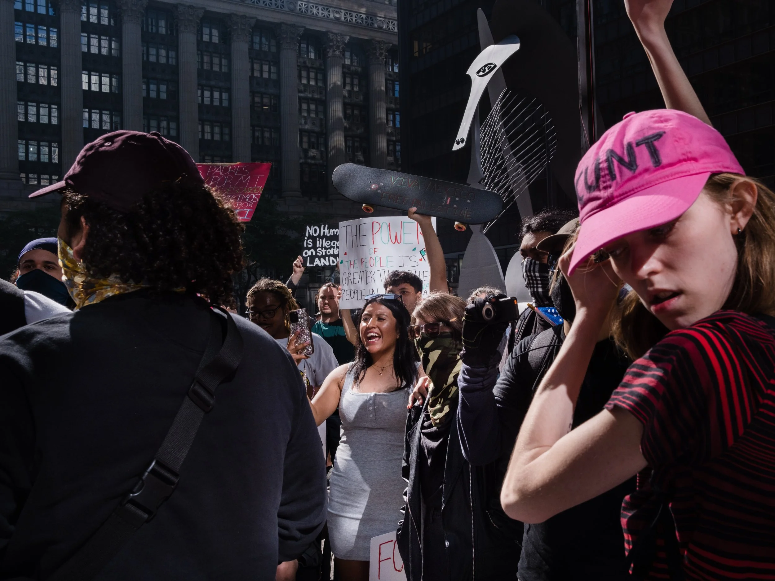 Crowd of protesters holding signs and banners in a demonstration in a city square. The signs include messages about human rights, power of the people, and illegal activities. The scene is busy with diverse individuals, some wearing masks, sunglasses,