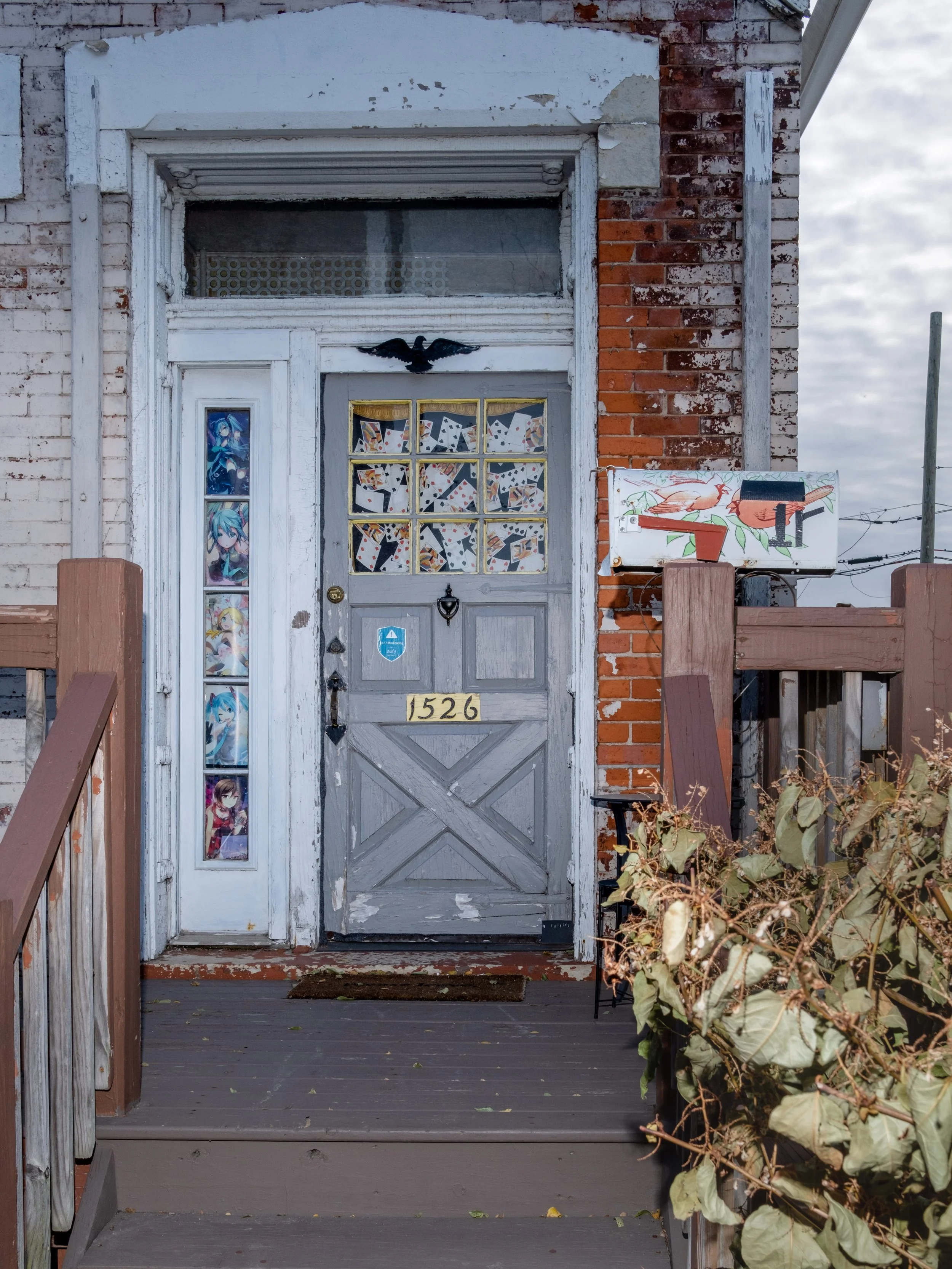 Abandoned house entrance with a weathered gray wooden door, decorated with various anime stickers, a black eagle sculpture above the door, and a side vertical column of anime posters. The house number 1526 is displayed on the door. There is a small p