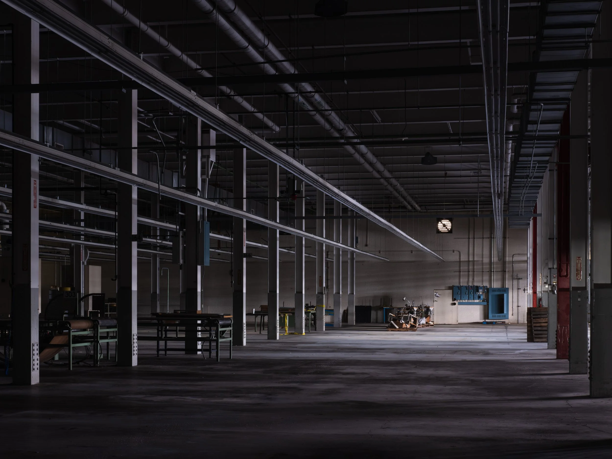 Empty industrial warehouse with metal pipes and equipment, dim lighting, and some machinery in the background.