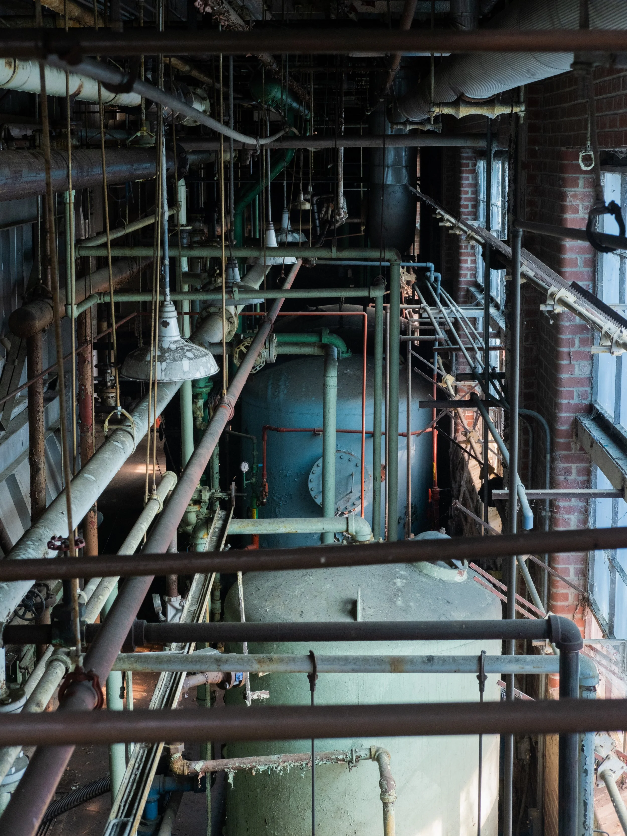 An industrial setting with numerous old pipes, a large blue tank, brick walls, and windows, showing a mechanical or boiler room.
