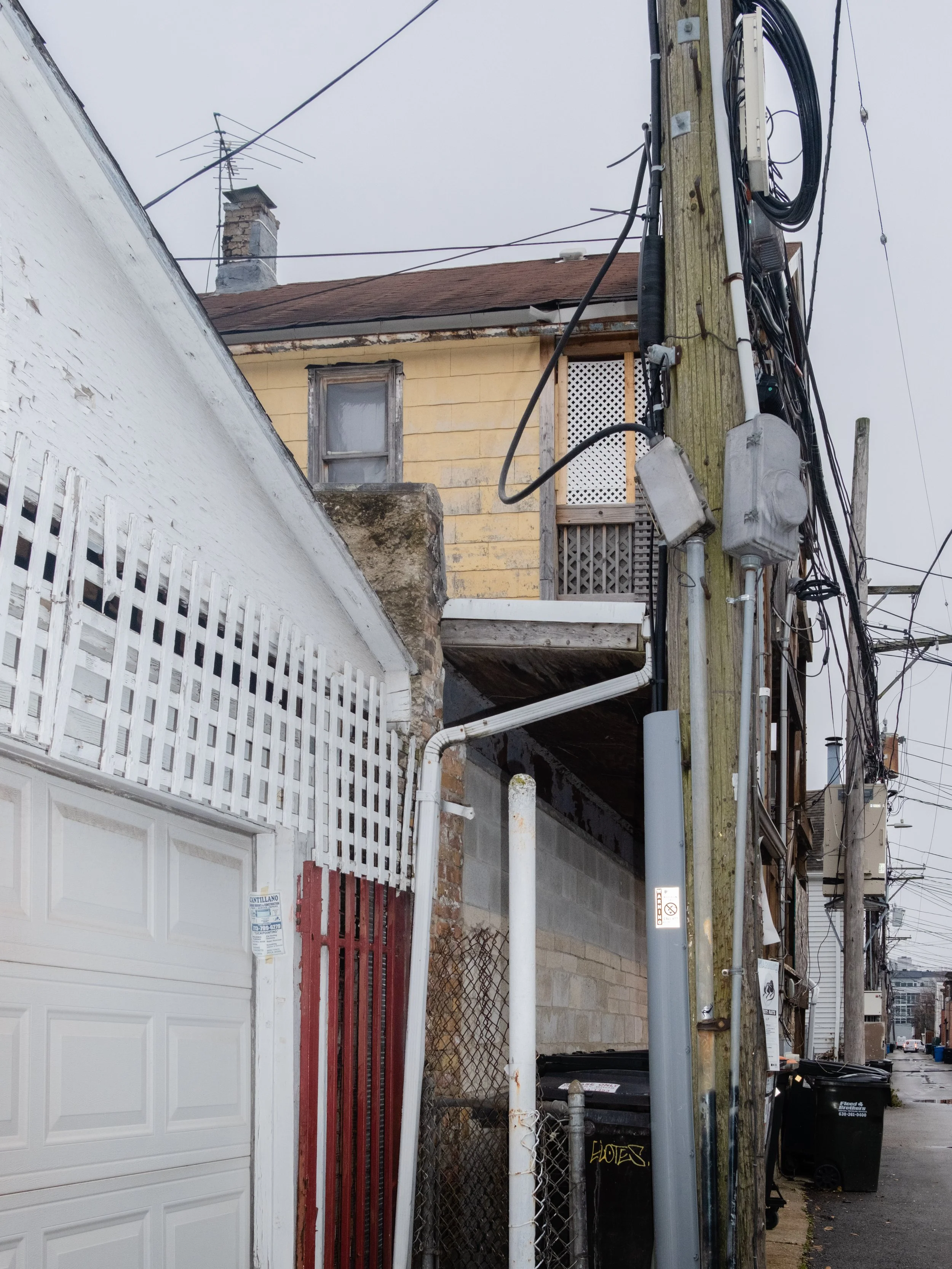 An alleyway scene with a utility pole featuring numerous wires, a yellow house with a second-story balcony, and a white garage door in the foreground.