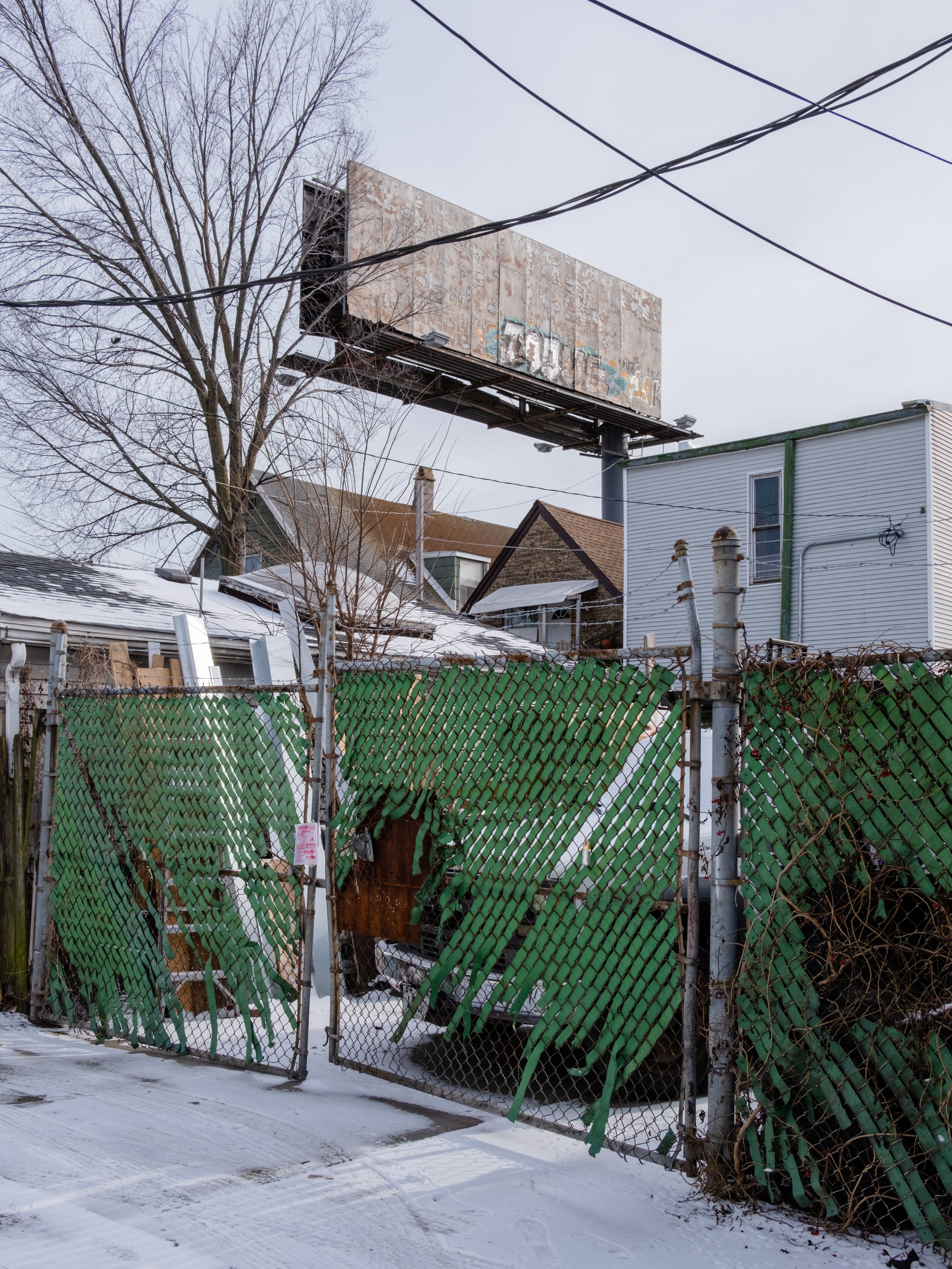 Urban alley with a snow-covered ground, a rusty chain-link fence with green plastic slats, a leafless tree, houses, power lines, and a large, weathered billboard structure above.