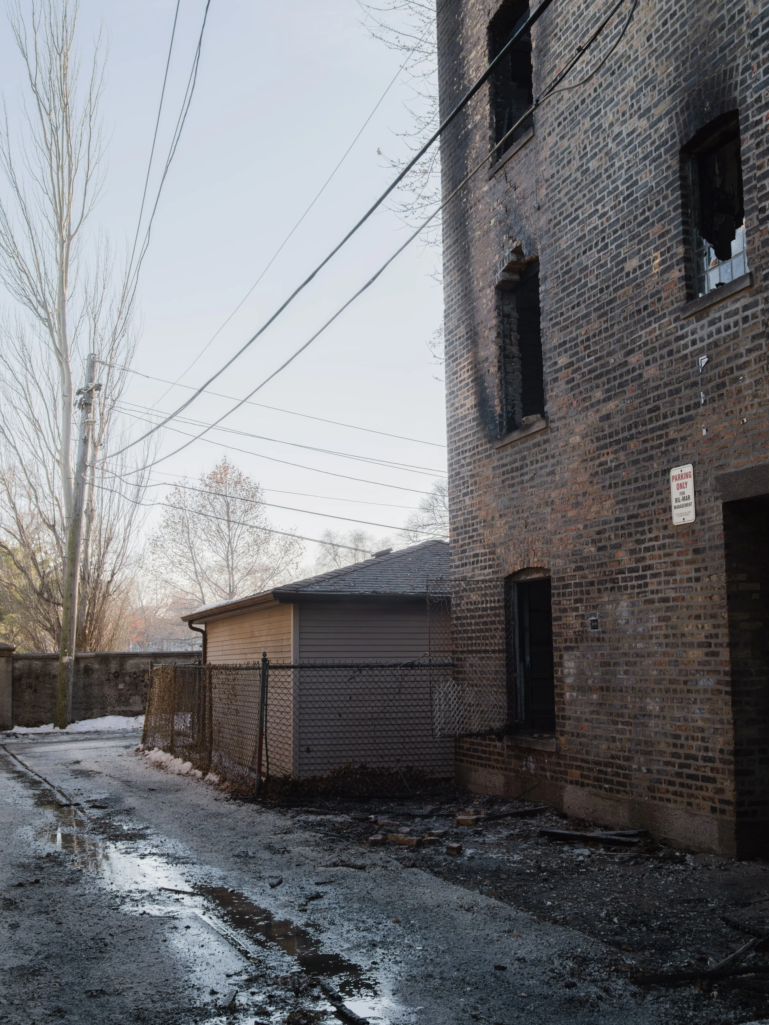 Back alley of a brick building with burned-out windows, damaged walls, and a wet, muddy ground with patches of snow, chain-link fence, and power lines overhead.