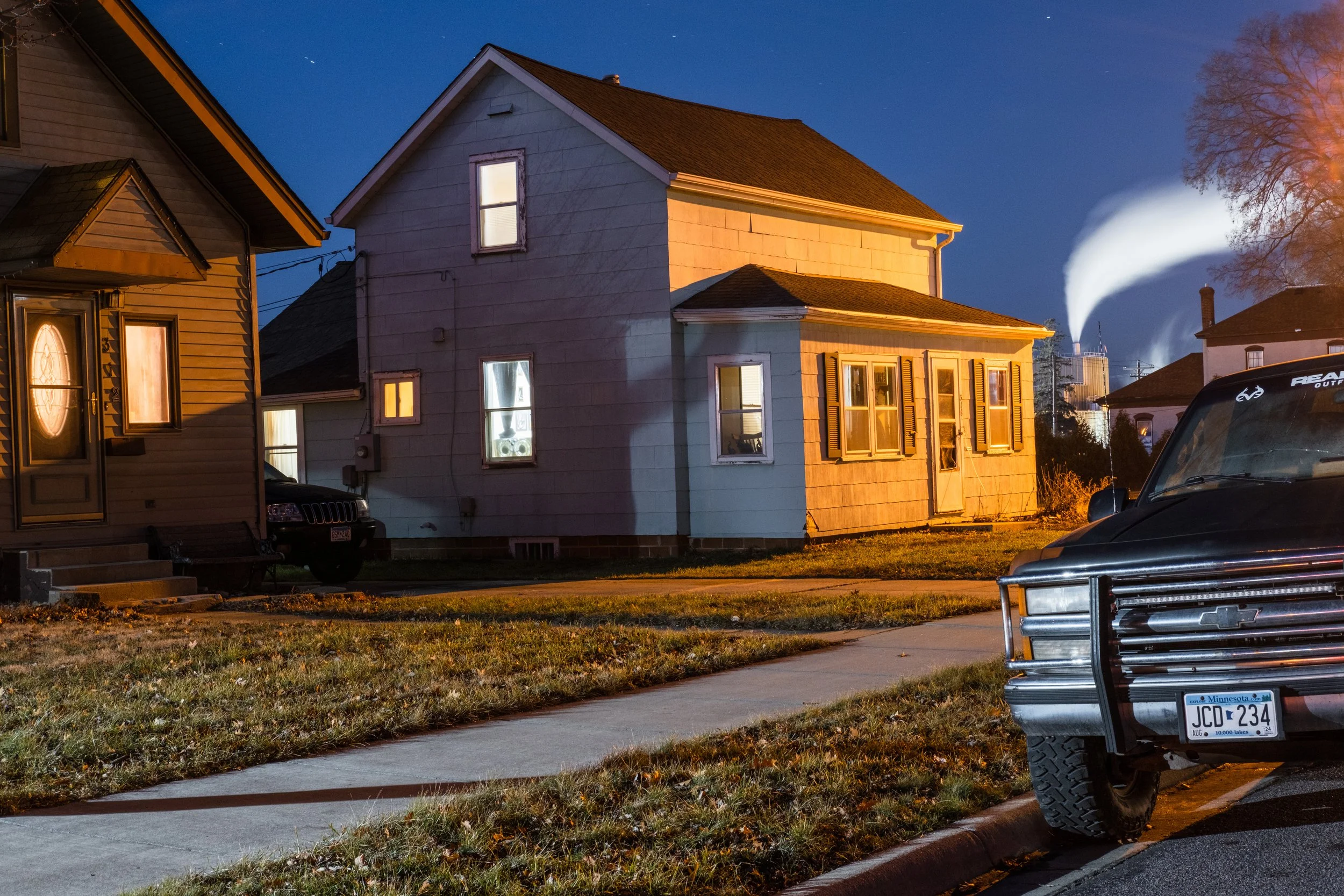 Nighttime scene of a suburban street with two houses, one painted in light blue and the other in brown with a front door illuminated. There are cars parked on the street, one black truck with a Minnesota license plate in the foreground, and a vehicle