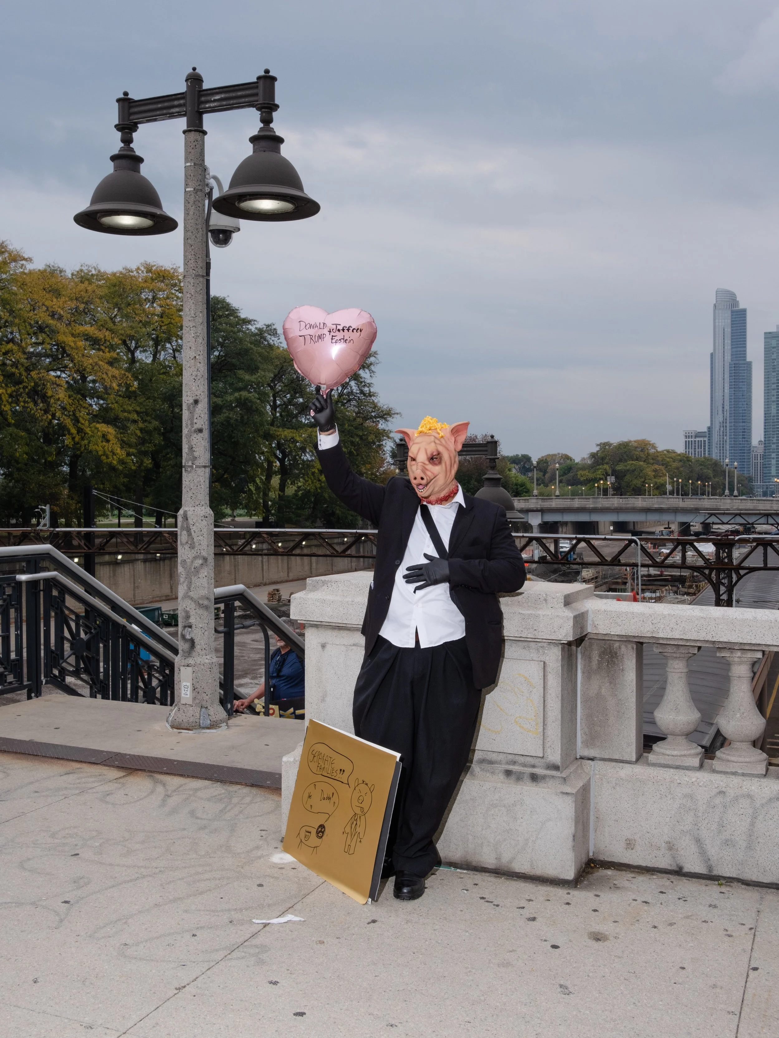 Person wearing a pig mask, black blazer, and holding a pink heart-shaped balloon with the words 'Donald J. Trump Forever' written on it, standing outdoors near a river with city skyline in the background.