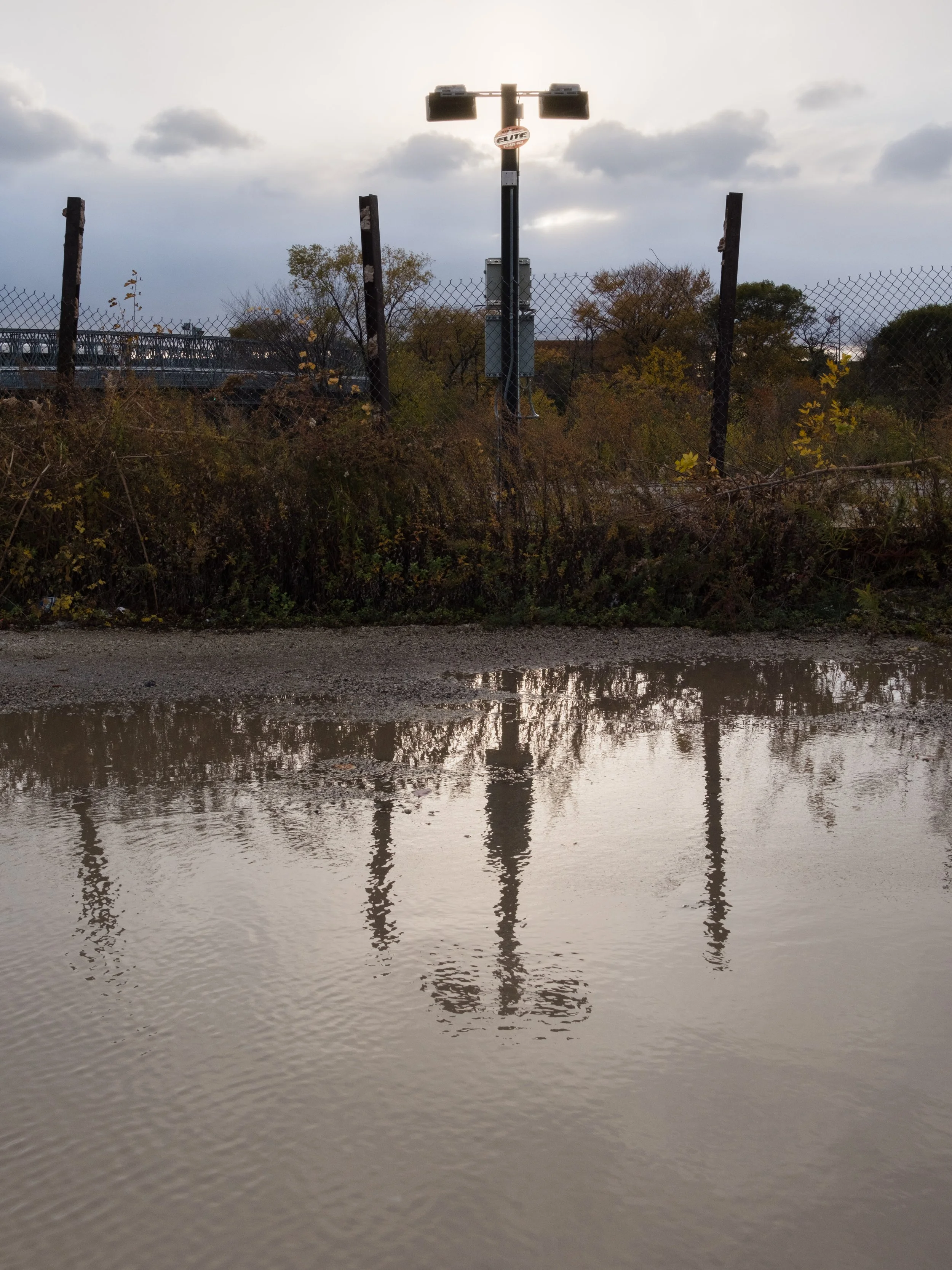 Flooded road with reflections of a light pole and fence, overcast sky with clouds.
