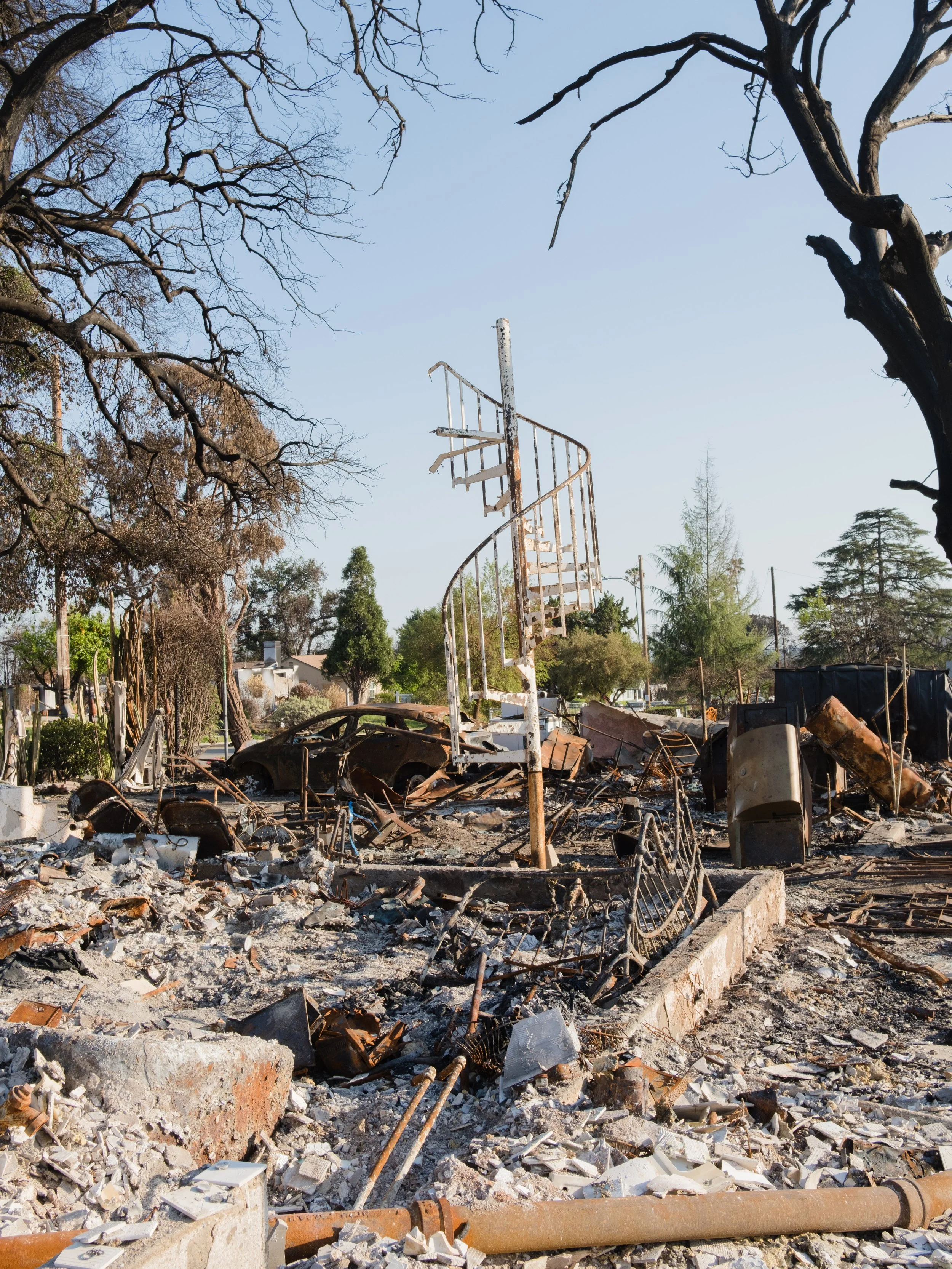 Burned-out neighborhood with twisted metal, charred trees, and debris, including a burnt car and a rusted spiral staircase.