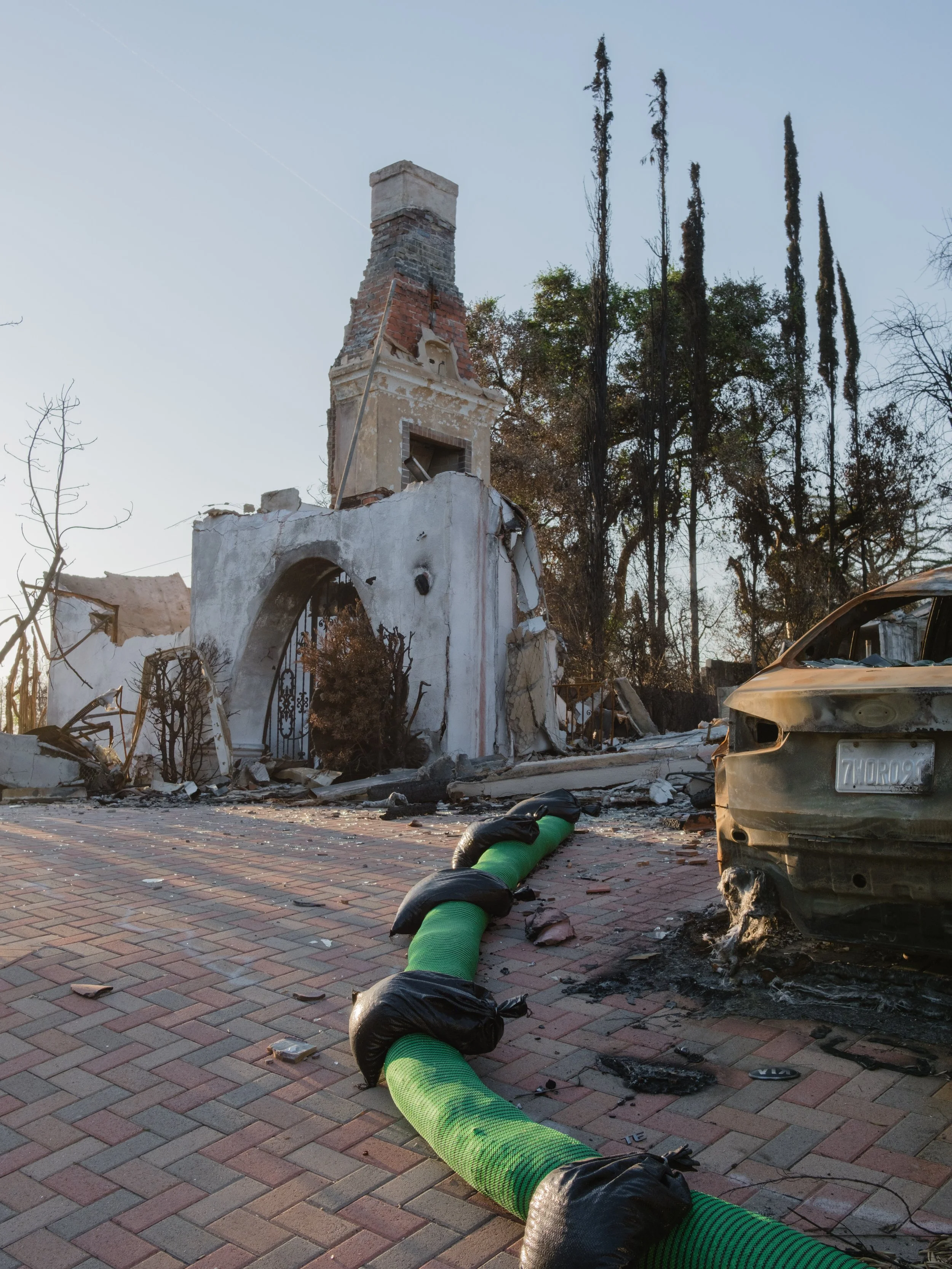 Damaged building with a chimney, debris, burnt-out vehicle, and a green hose on a brick-paved street.