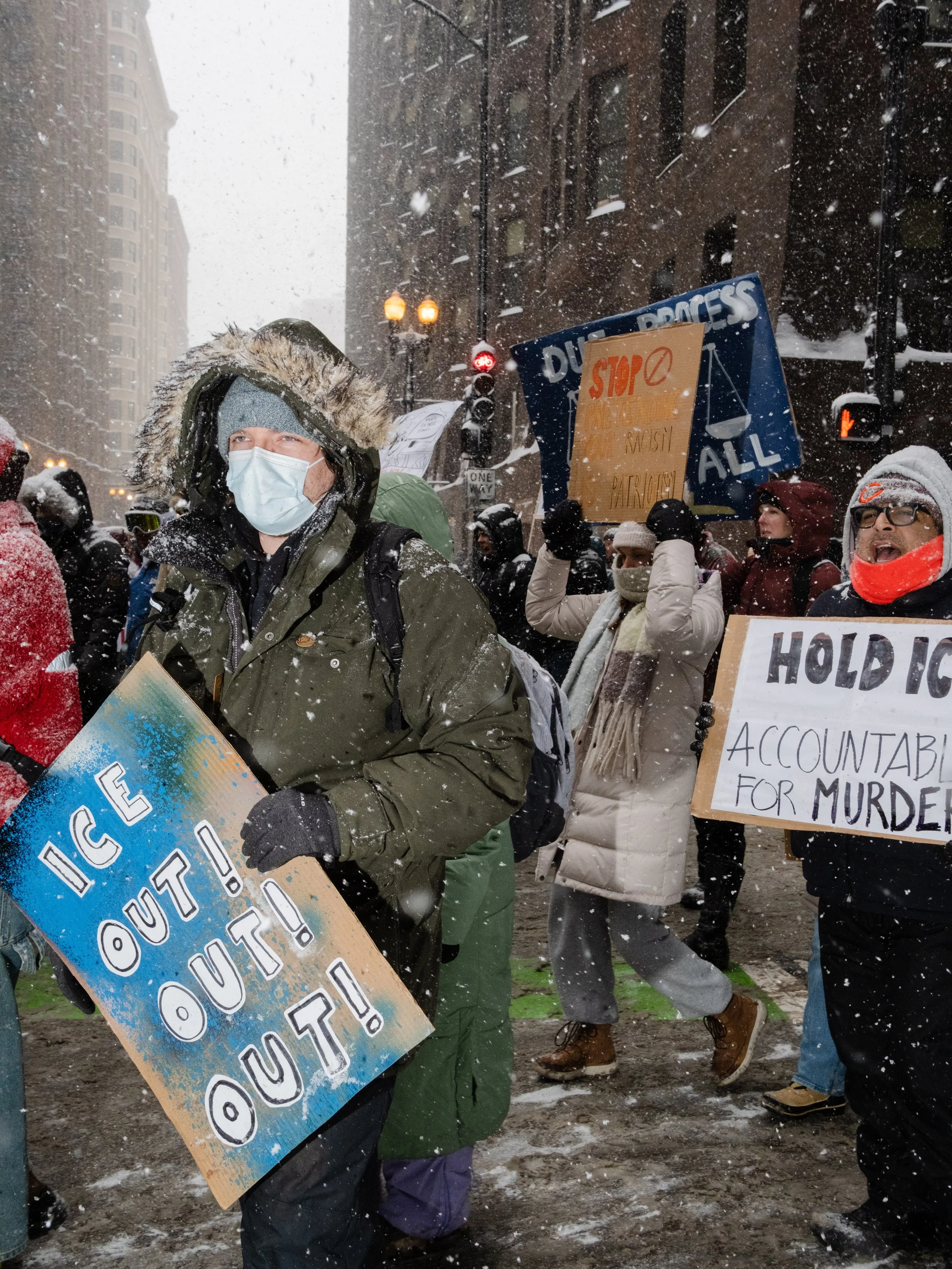 Protesters wearing winter clothing and face masks holding signs during a snowstorm in an urban setting. Signs include messages like 'ICE OUT!' and calls for accountability and stopping harmful policies.