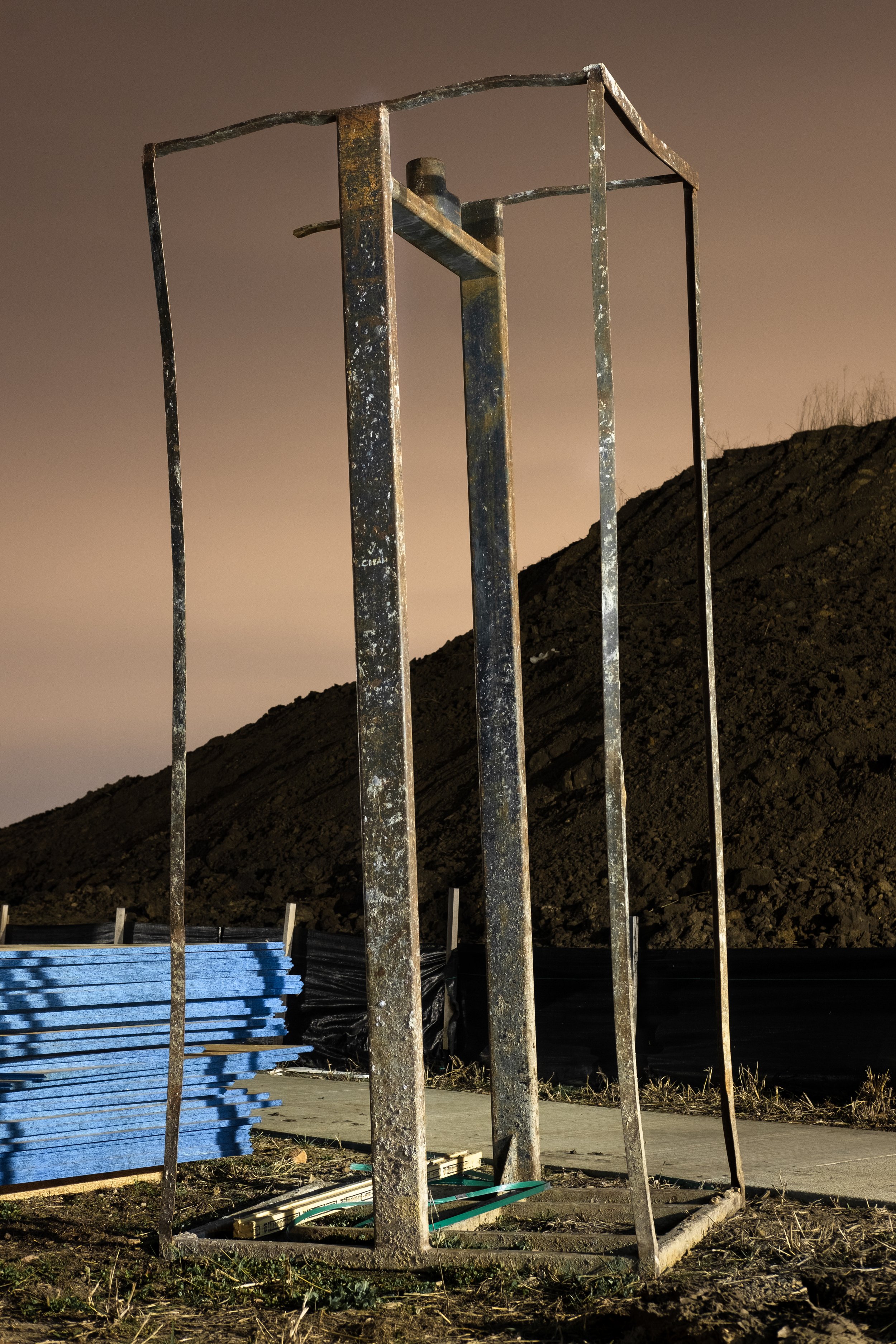 An old, rusted metal frame standing outdoors on a dirt surface near a sidewalk, with a hill and a cloudy sky in the background.