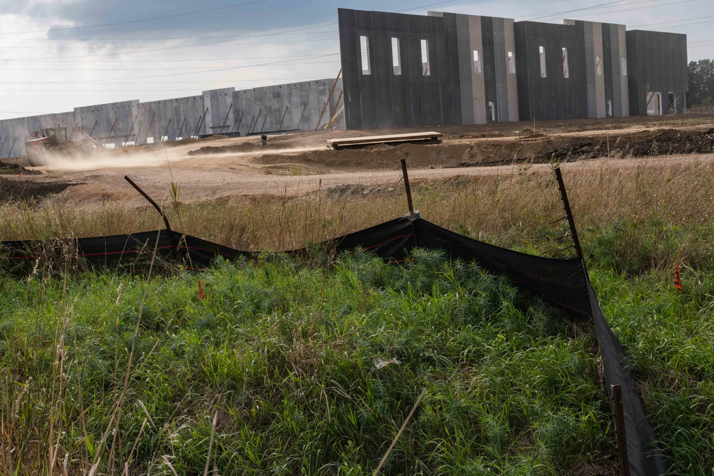 Construction site with a modern building under construction in the background, dust rising from the dirt ground, and black safety fence in the foreground.