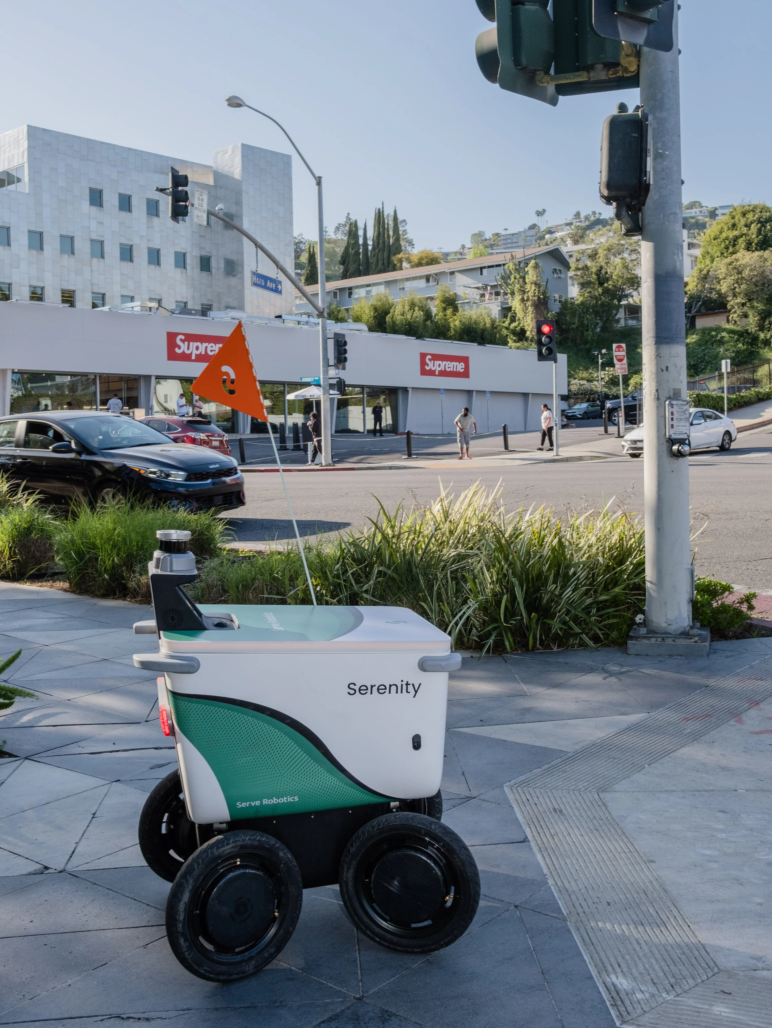 A white robot with four wheels parked on the sidewalk near a street corner. The robot has the words 'Serenity' and 'Serve Robotics' on it, with an orange flag on a pole attached to it. In the background, there is a street with cars, pedestrians, traf