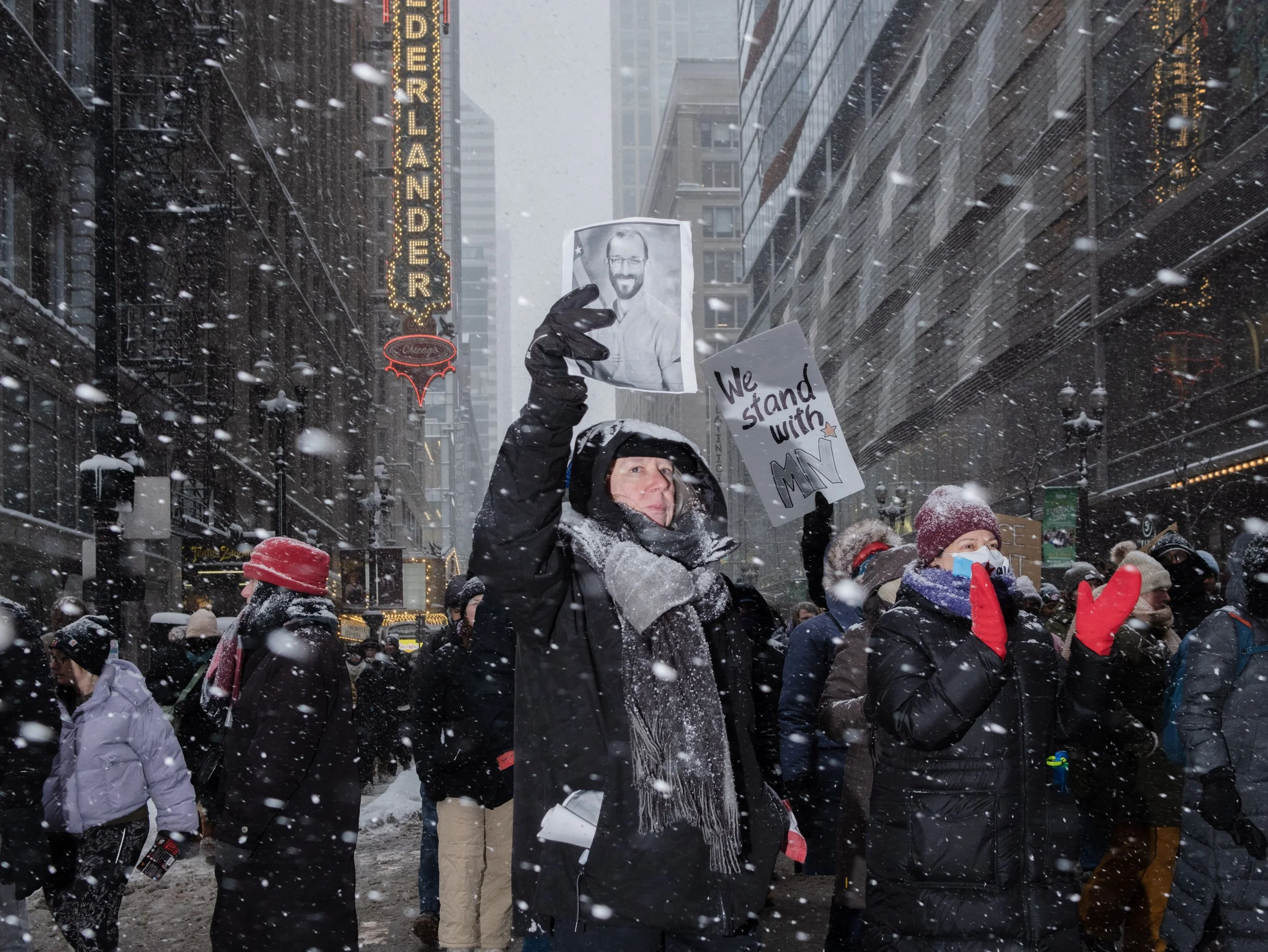 A person in winter clothing holding a sign with a photo of a man, and another sign that says "We stand with MN," during a snowstorm in a city street with other protesters around.