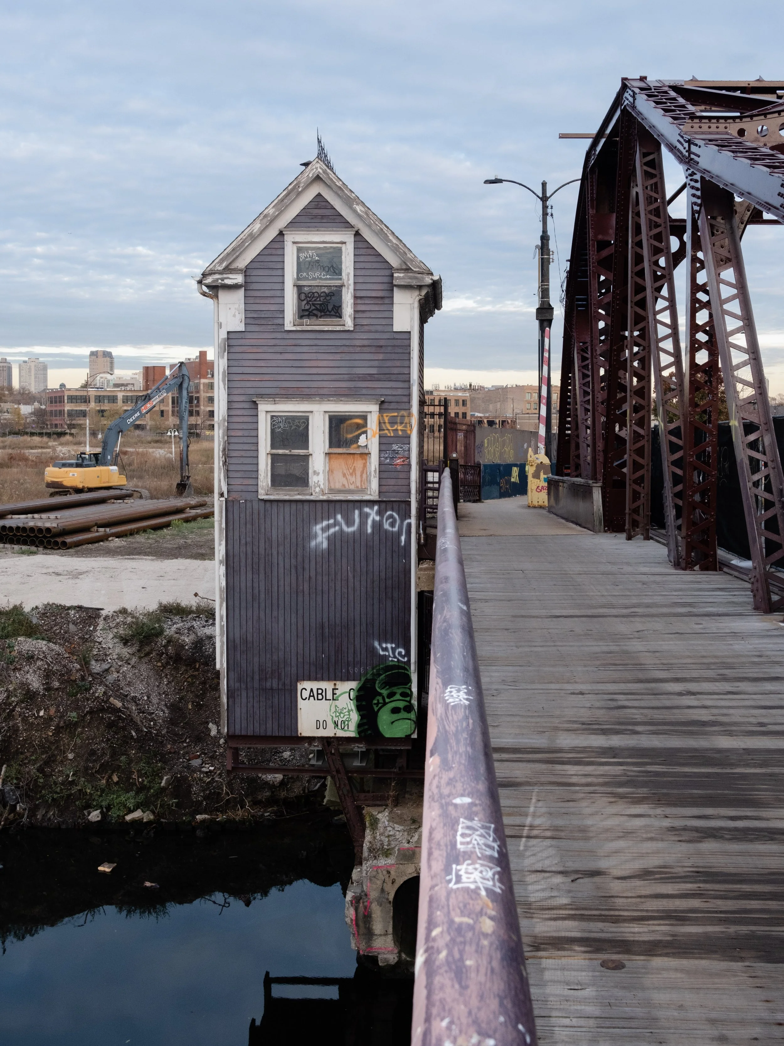 A small, weathered house with boarded-up windows and graffiti, situated near a bridge with a rusted metal structure. An excavator and industrial pipes are visible in the background under a cloudy sky.