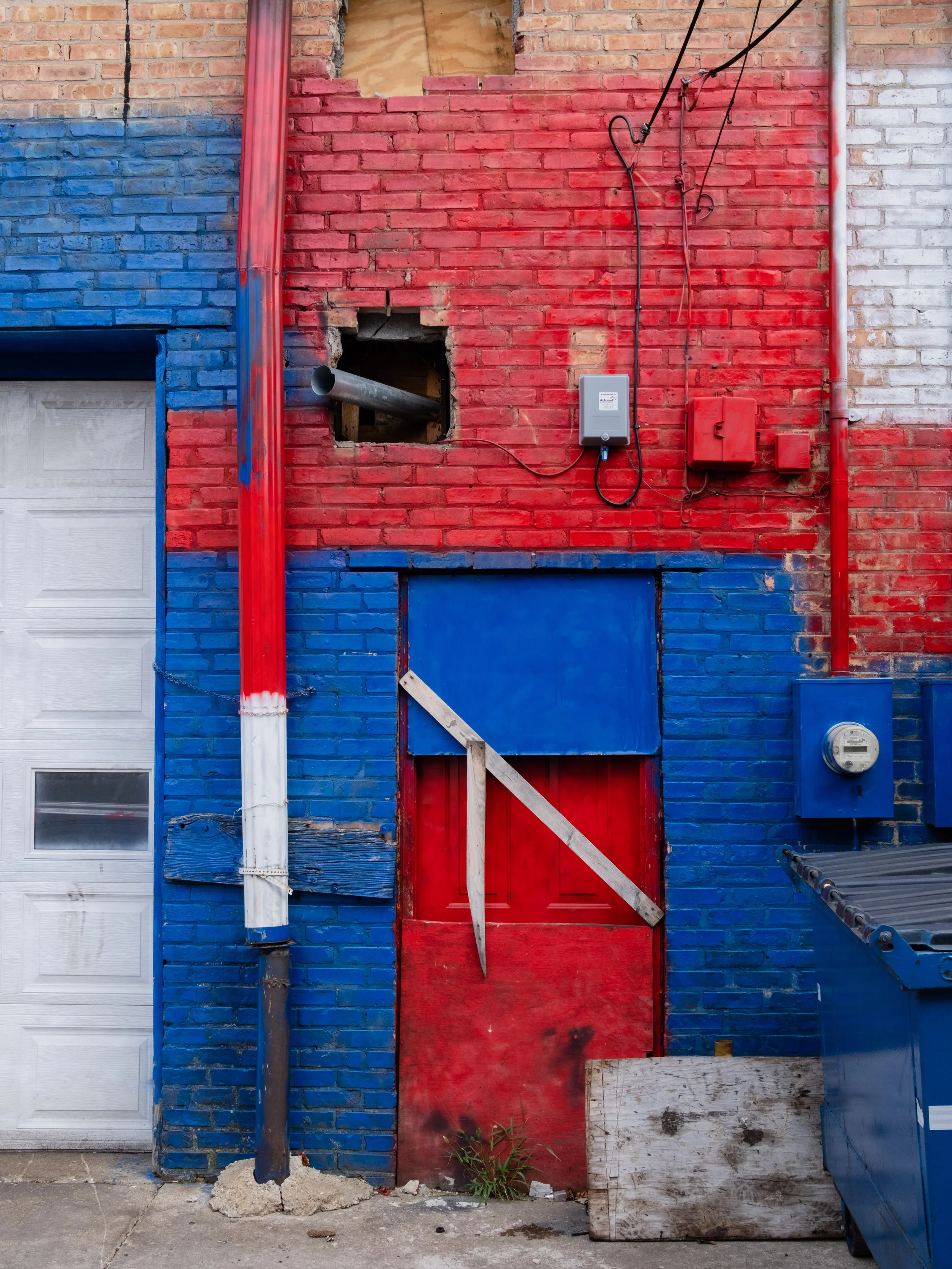 The wall of a building painted with red and blue sections. There is a white garage door on the left, a hole in the brick wall with a pipe sticking out, and a makeshift red and blue door with a wood brace in the middle. Various electrical meters and w
