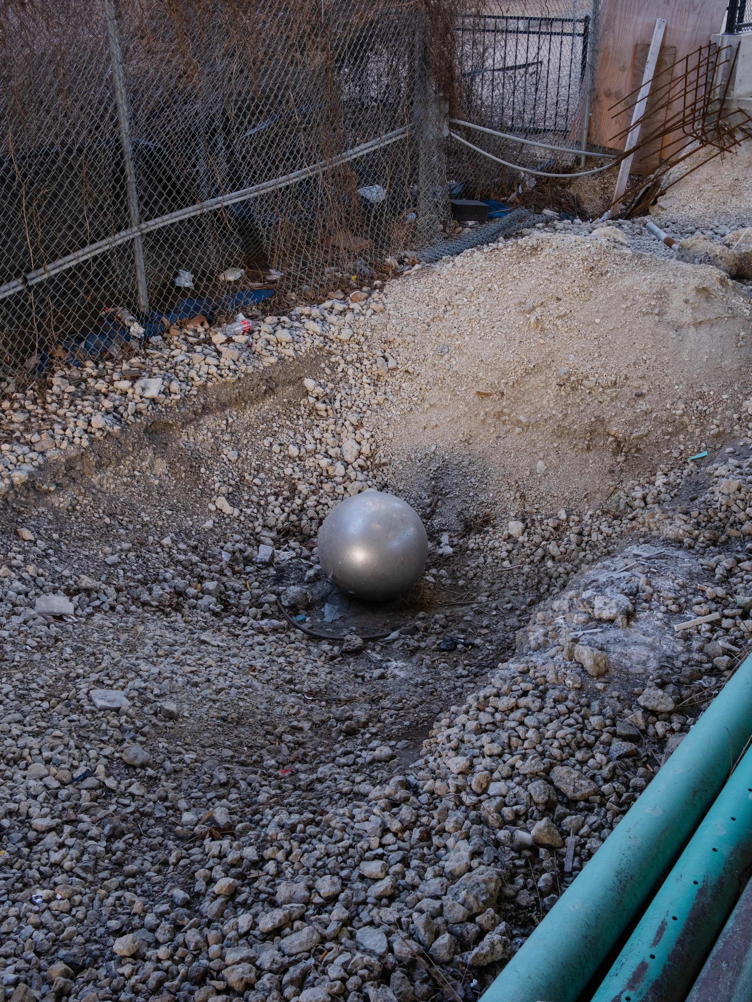 A residential backyard construction site with a large metallic ball, a dug hole, gravel ground, fencing, and some pipes.