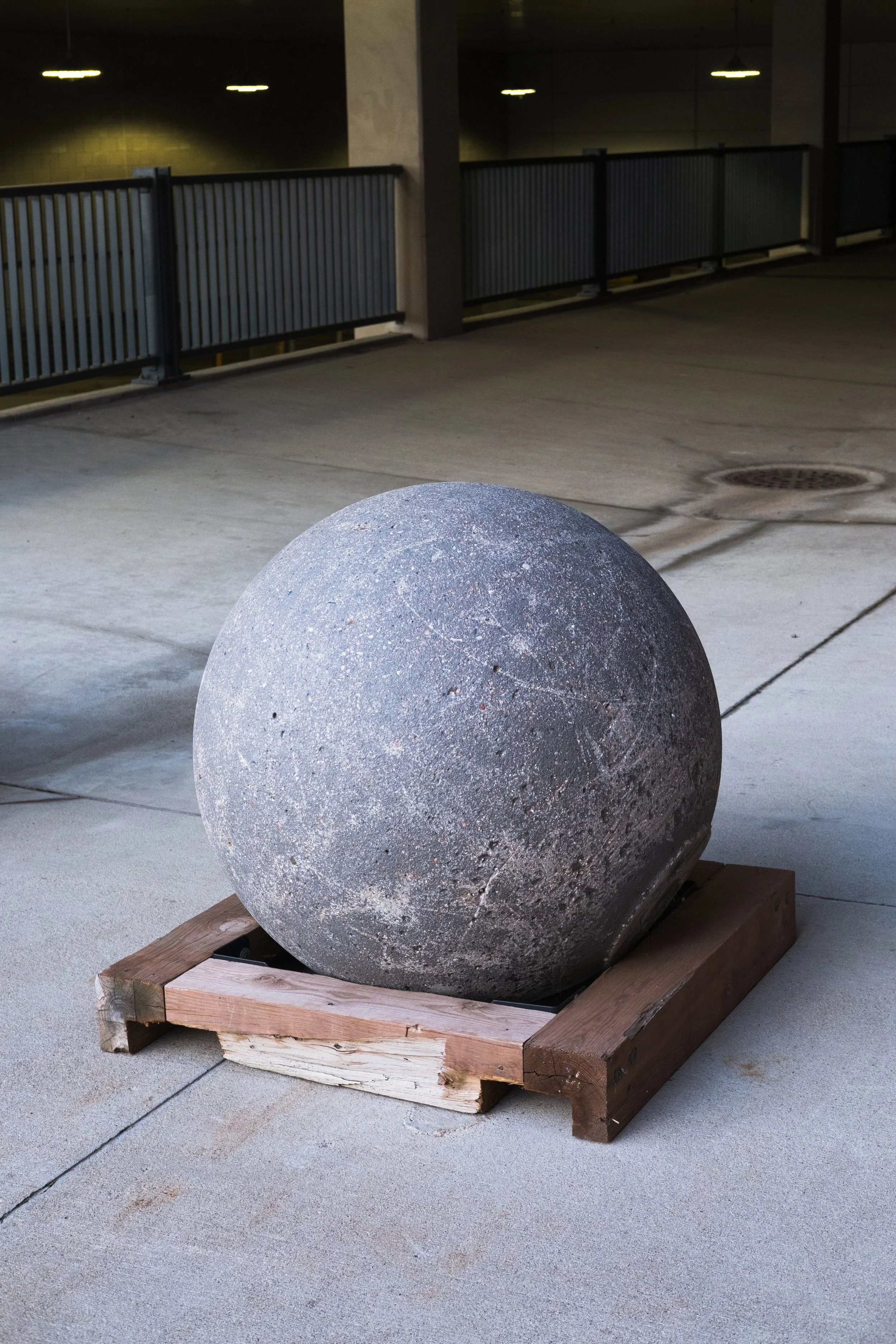 A large concrete ball resting on a wooden frame in an outdoor parking garage at night.