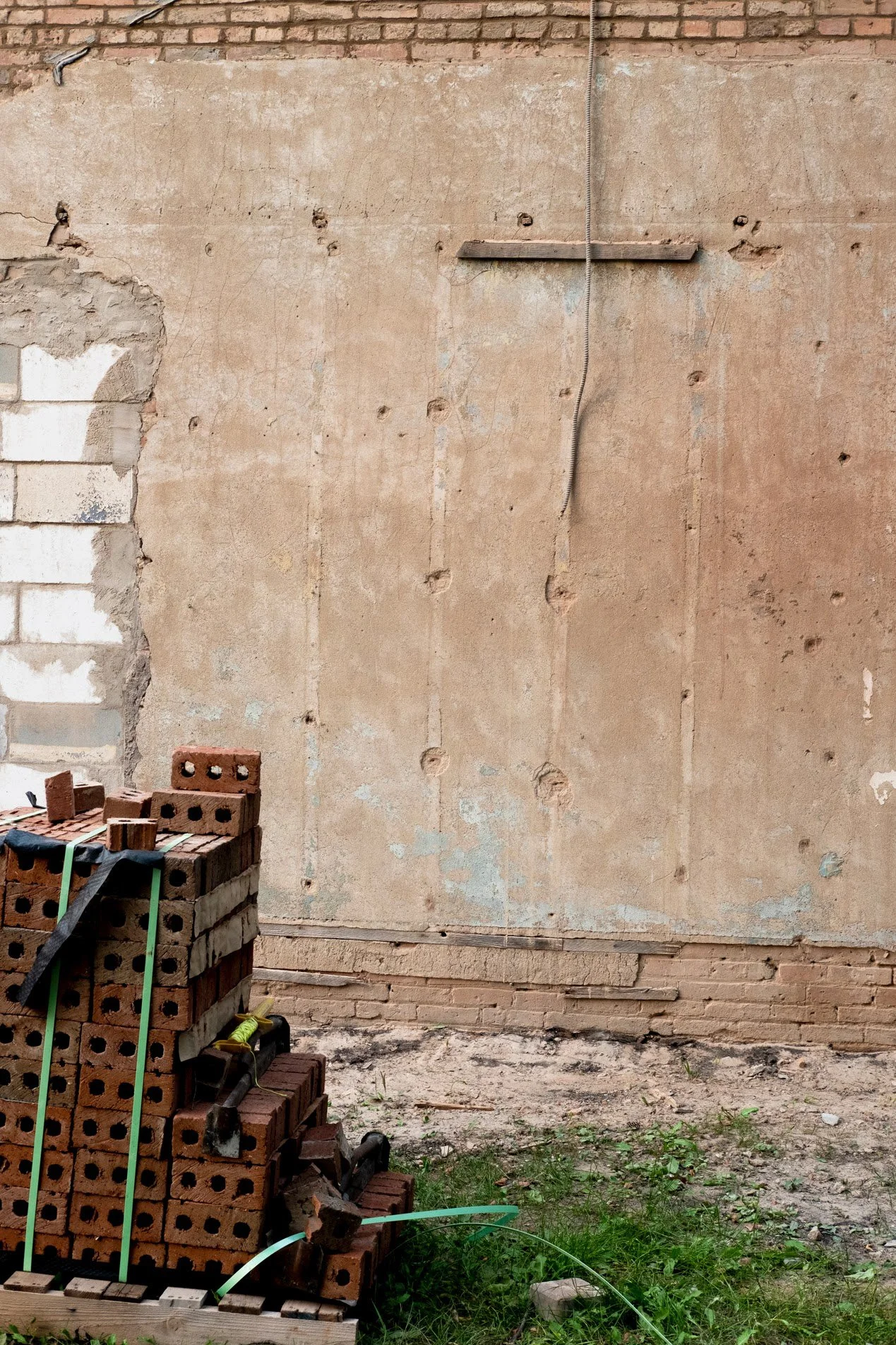 A stack of red bricks tied with green straps, placed on a wooden pallet outside a building with an unfinished wall, some bricks, a coil of wire, and patches of grass and dirt.
