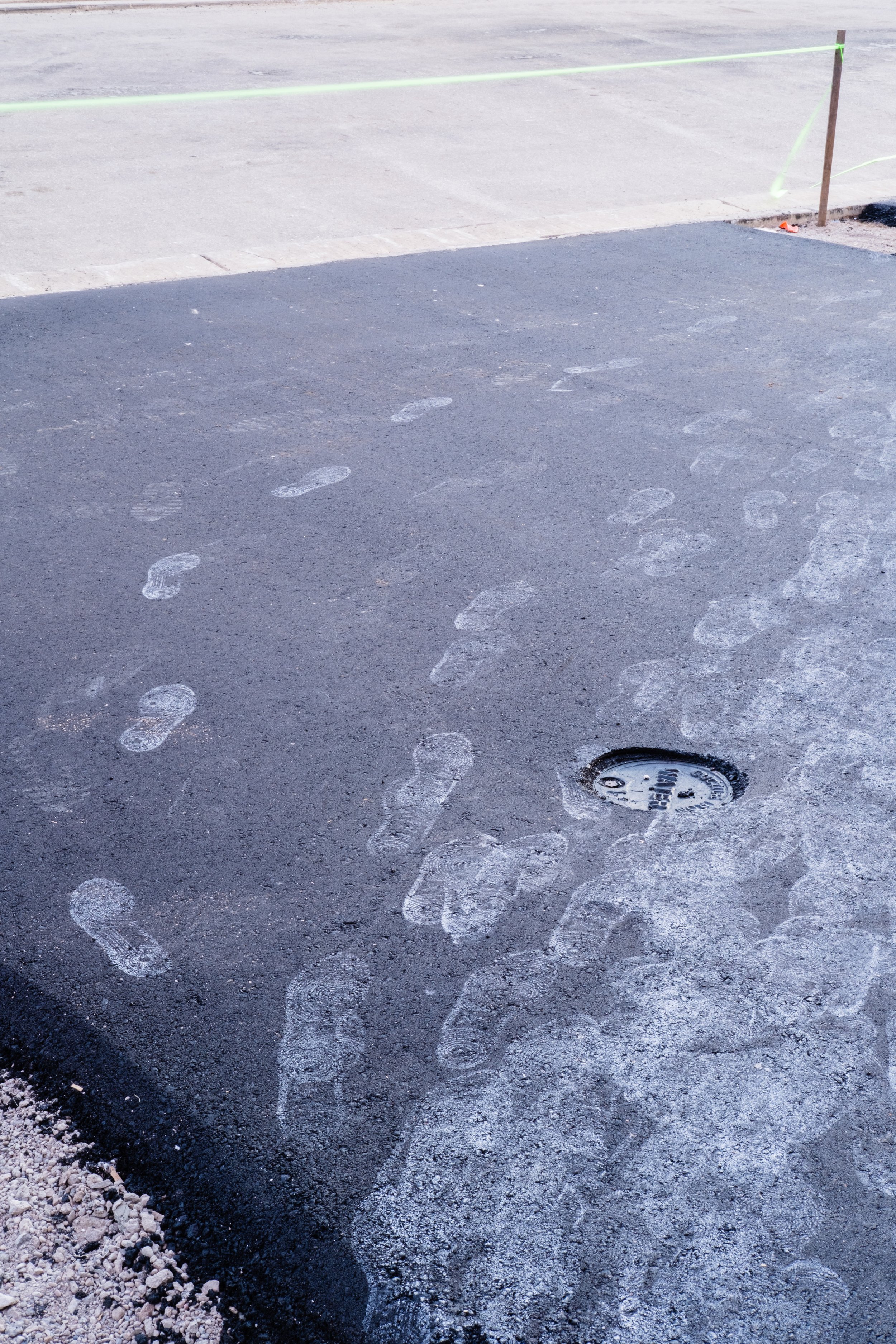 Footprints in fresh asphalt leading to a manhole cover outside, with construction barrier tape in the background.