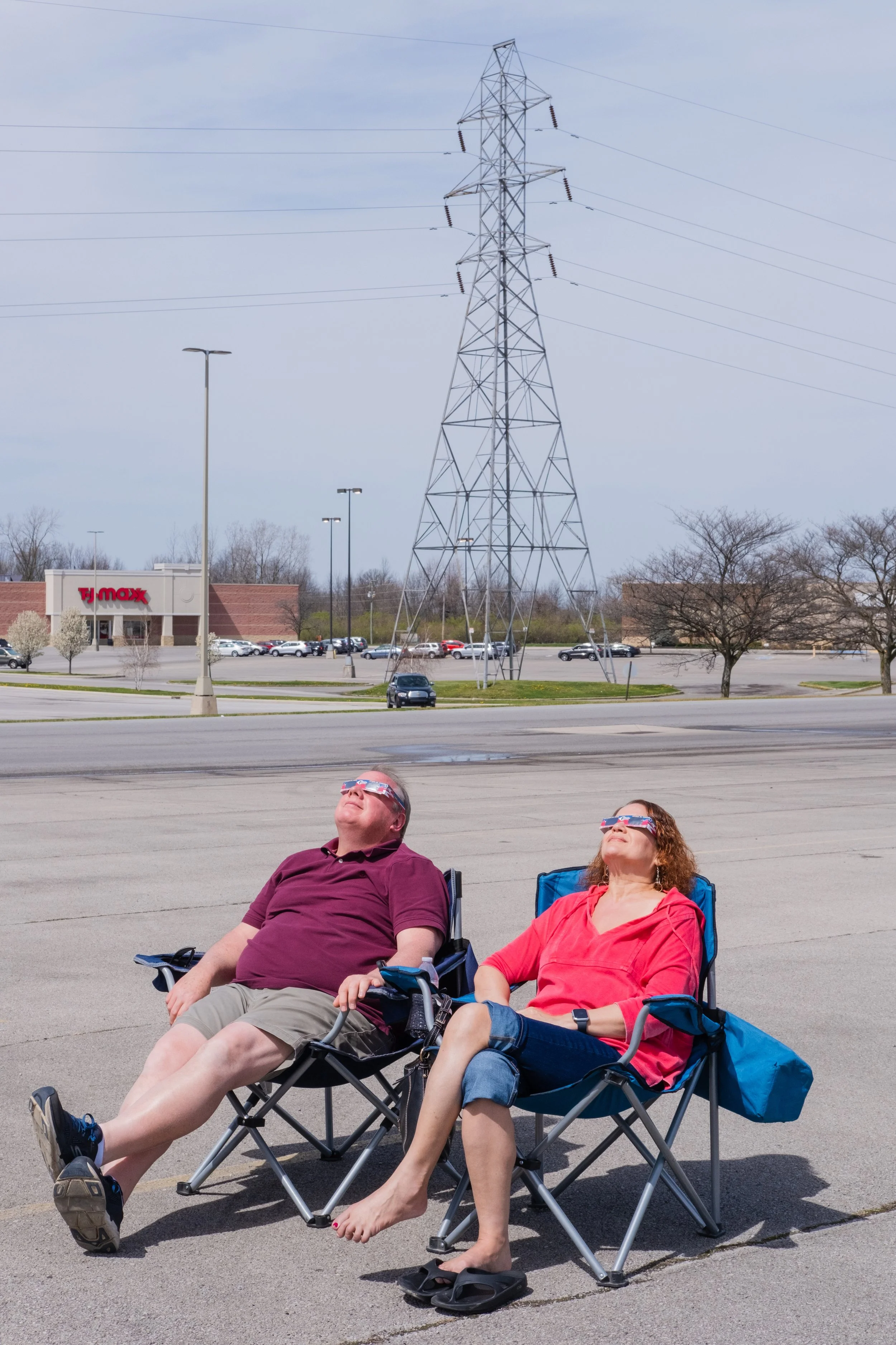 A man and woman relaxing in portable chairs outdoors during sunny weather, wearing 3D glasses, sitting in a parking lot near power lines and a TJ Maxx store.