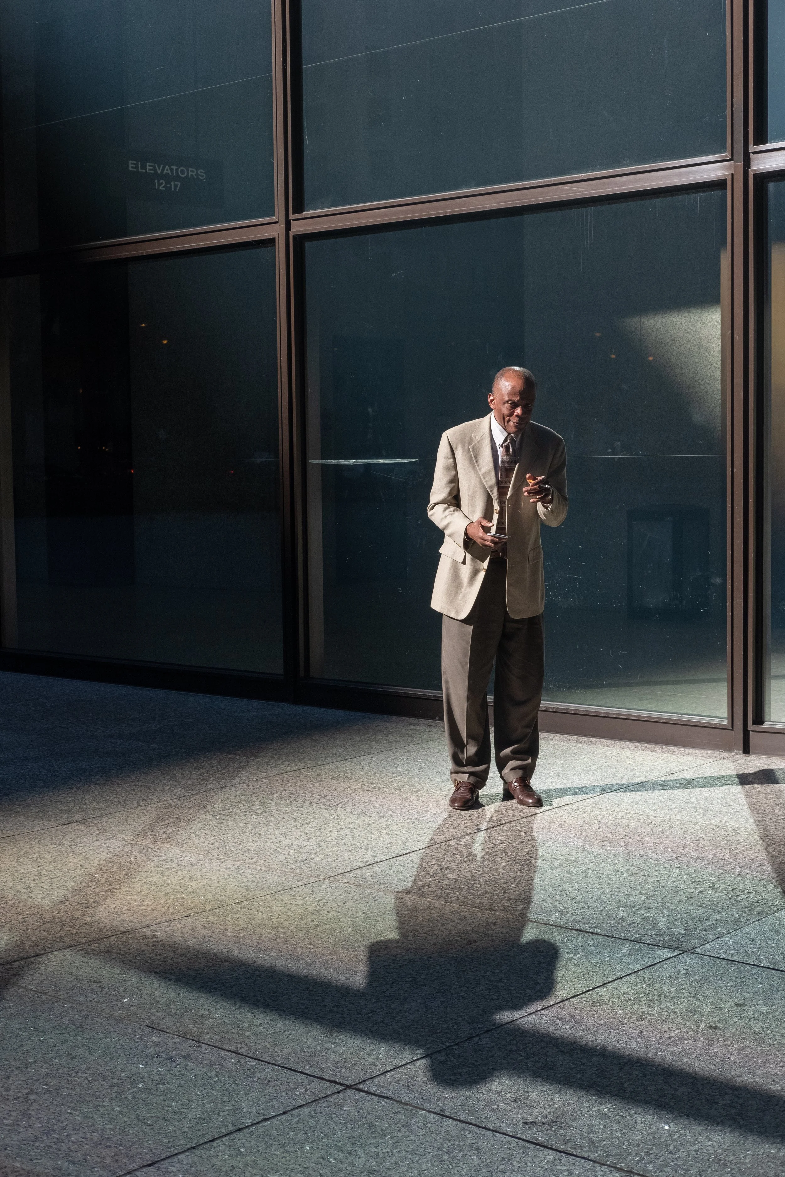An elderly man in a beige suit and brown shoes standing outside near a glass building, looking down at his phone, with a shadow cast on the pavement.